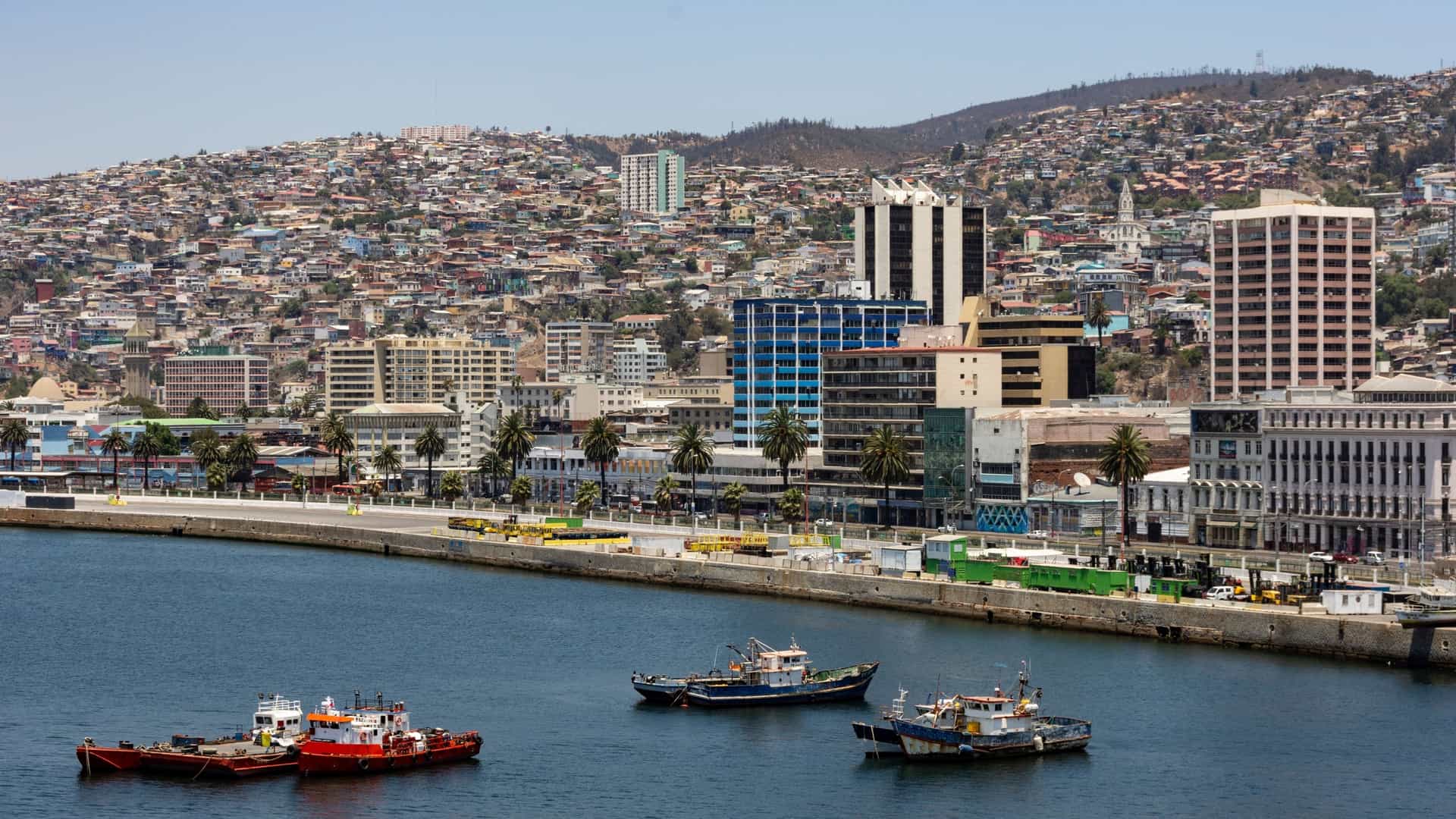A wide, high angle panoramic view of the port city of Valparaíso, Chile. The image captures the dense, colorful cluster of houses built into the steep hills (cerros) overlooking the flat city center, the industrial harbor, and the waterfront, with the Pacific Ocean stretching out to the horizon under a bright sky. The scene highlights the unique, amphitheater like urban layout of this UNESCO World Heritage Site.