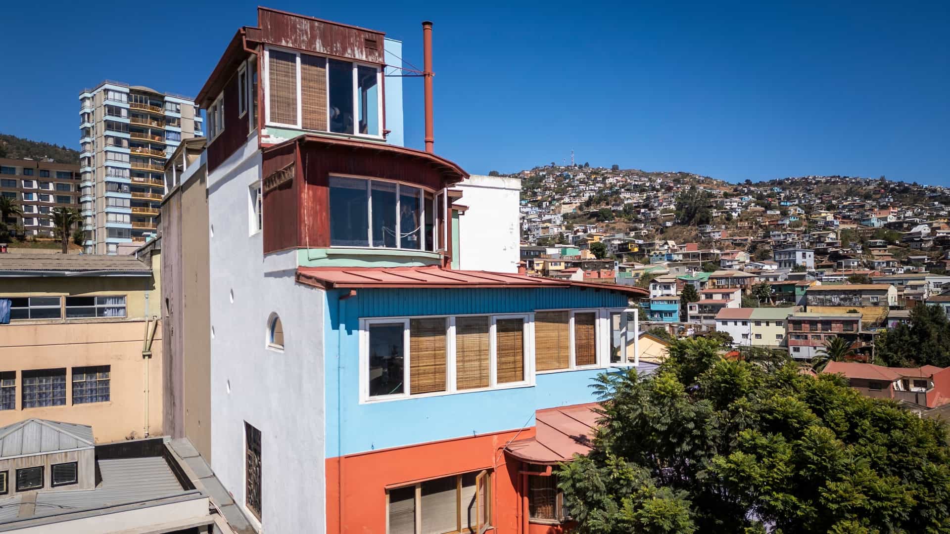 A beautiful aerial view of La Sebastiana, the distinctive, multi story house museum of poet Pablo Neruda, perched on Cerro Florida in Valparaíso, Chile. The unique, brightly colored architecture of the house stands prominently among the surrounding hillside residences, offering a sweeping panorama of the densely packed UNESCO World Heritage city, the harbor, and the Pacific Ocean.