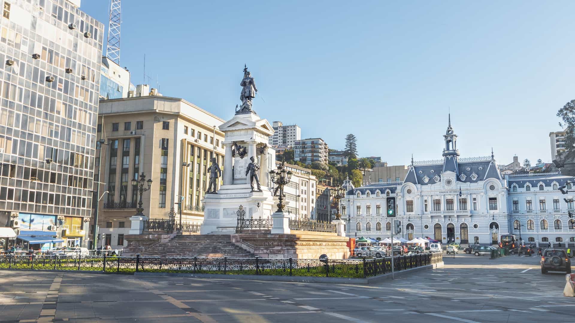 A photograph of the iconic Plaza Sotomayor in Valparaíso, Chile, centered on the neoclassical Edificio Armada de Chile (Chilean Navy Headquarters). The ornate blue and white building, featuring a prominent clock and mansard roof, forms the backdrop for the square, which is the site of the Monument to the Heroes of Iquique, a central column honoring naval heroes.