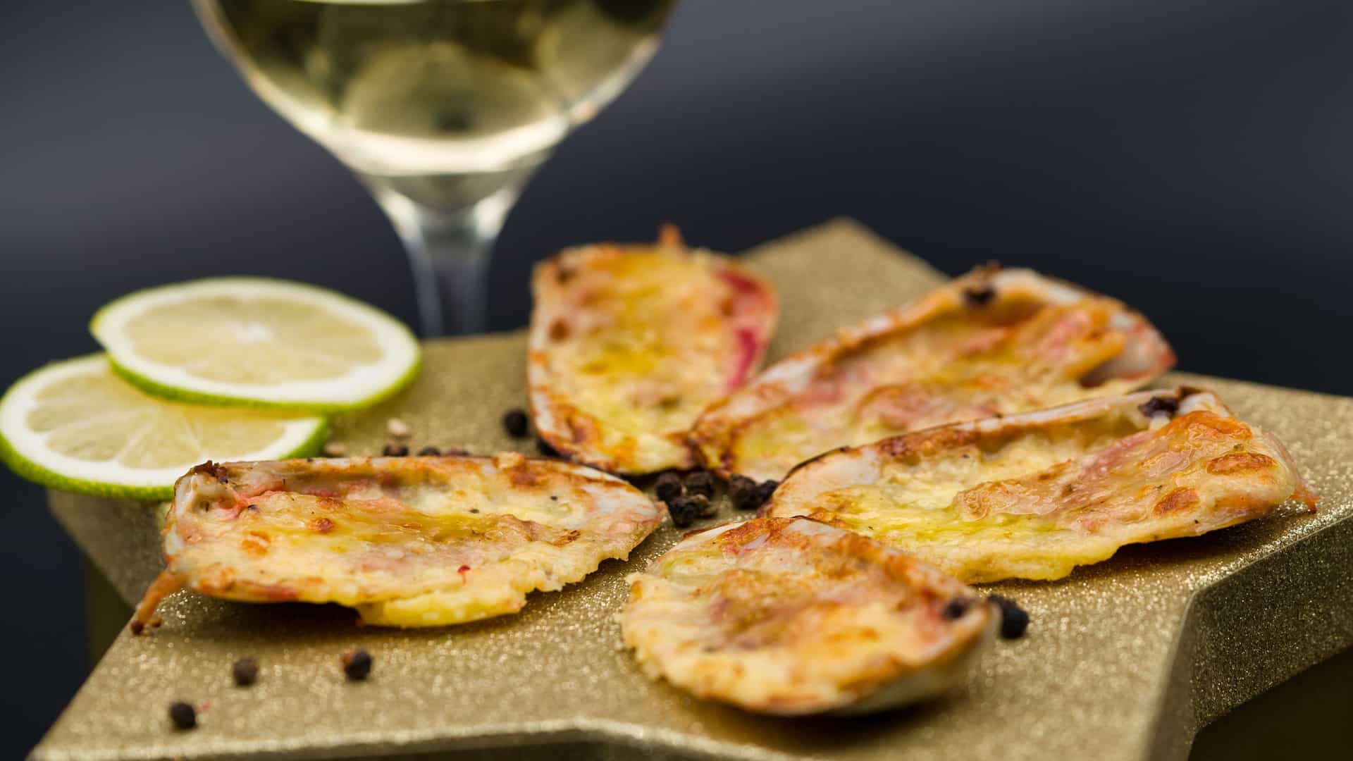 A close up view of Machas a la Parmesana, a traditional Chilean seafood appetizer. The image shows several razor clams (machas) nestled in their shells, topped with melted and gratinéed golden Parmesan cheese. This savory dish is a staple of coastal Chilean dining, often enjoyed in restaurants overlooking the waterfront.