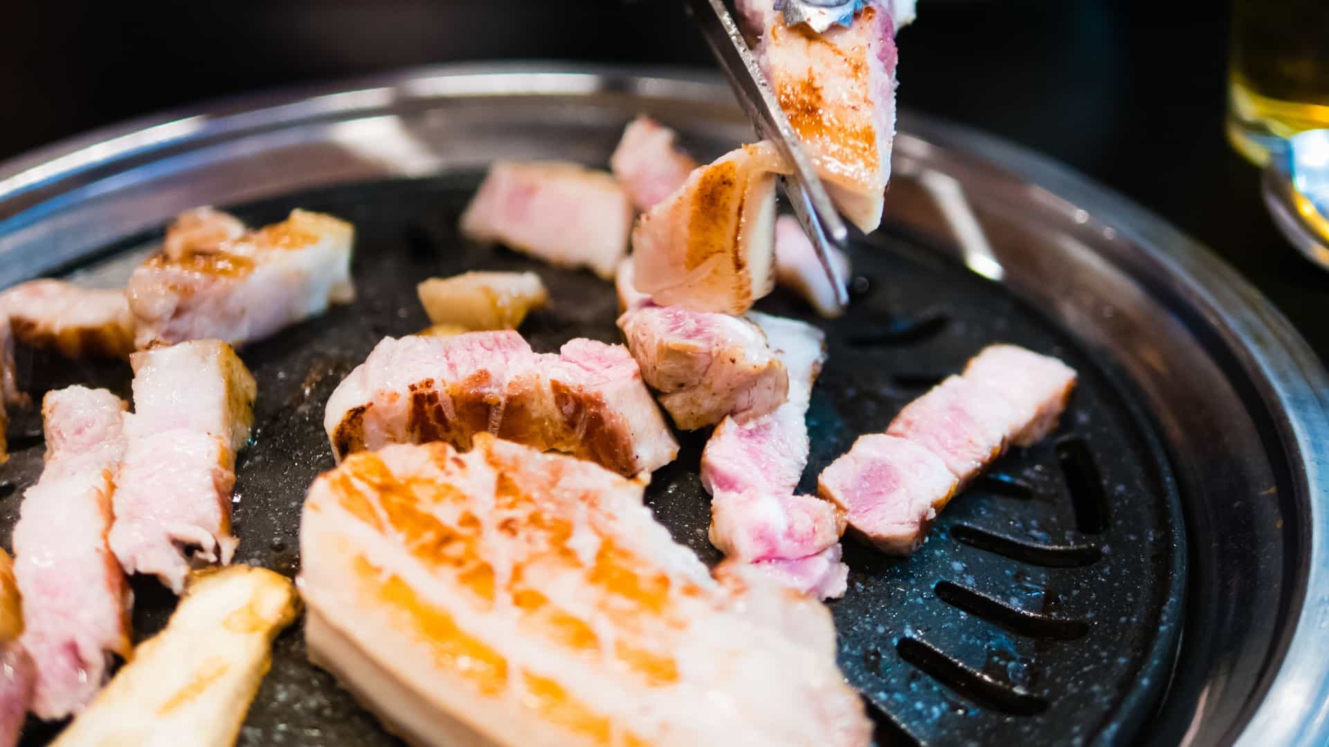 Slices of raw, thick cut pork belly, known as Samgyupsal, are laid out on a hot, slanted Korean style grill pan, surrounded by various Korean side dishes, including kimchi, garlic, and fresh vegetables, ready for a table side barbecue.