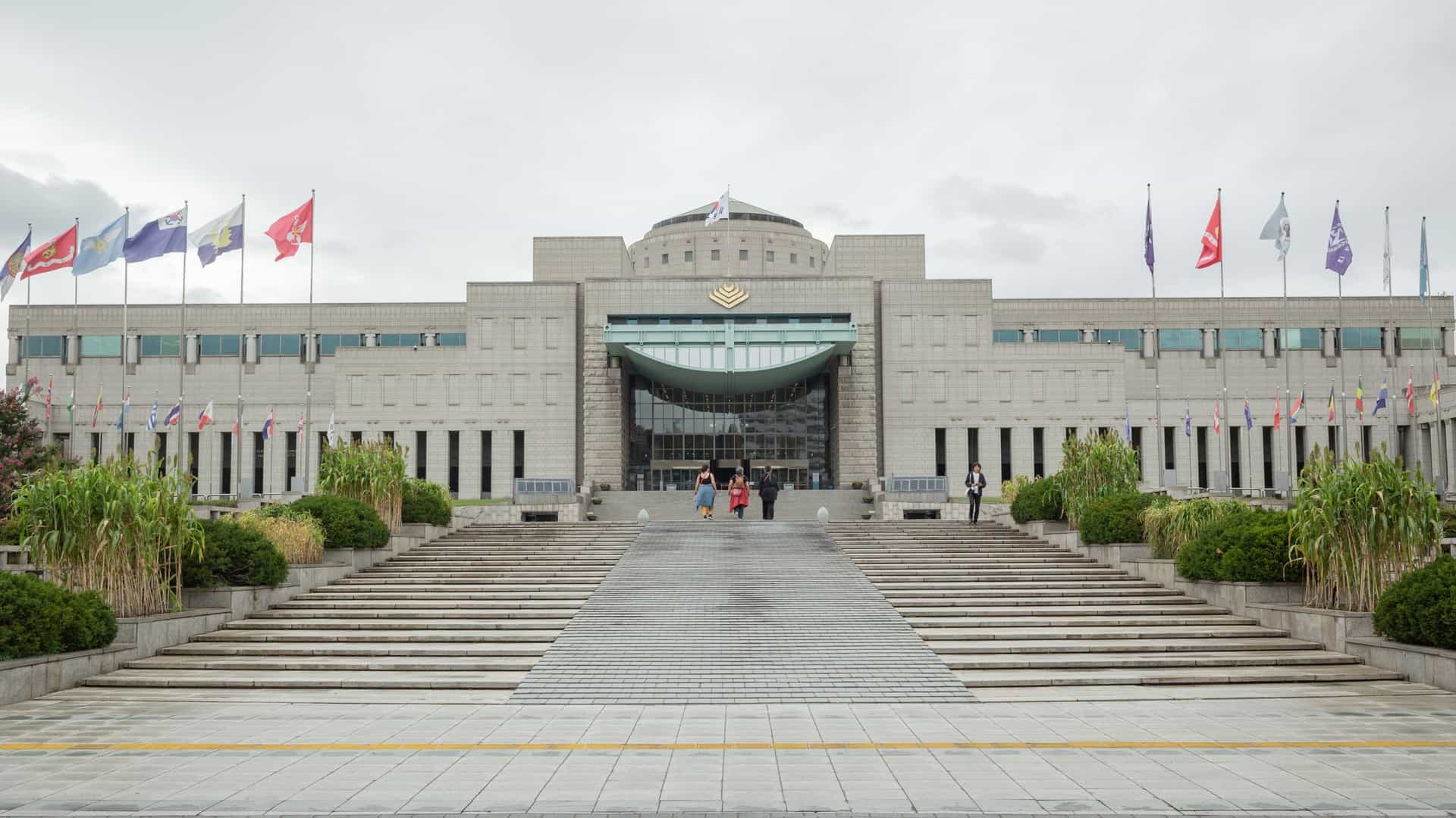 The exterior of the War Memorial of Korea in Seoul, featuring the grand, modern museum building in the background. In the foreground are the two towering, streamlined columns of the Korean War Monument, flanked by a group of commemorative statues in the main plaza.