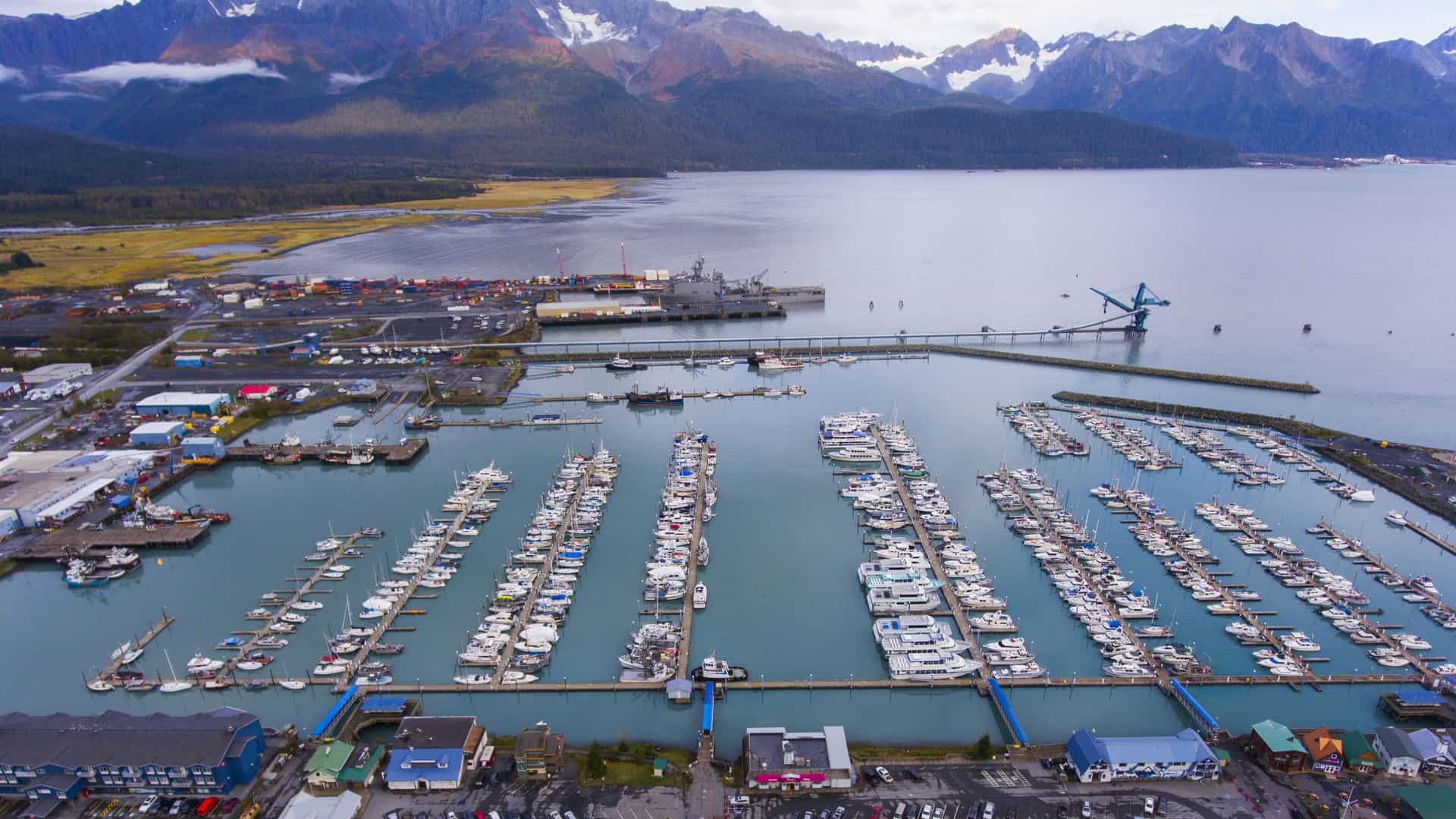An aerial view of the Seward Boat Harbor and waterfront in Alaska. The harbor is densely packed with rows of commercial fishing boats and recreational vessels docked in the turquoise water of Resurrection Bay. The city's waterfront structures line the shore, set against a dramatic backdrop of large, rugged mountains from the Kenai Peninsula.