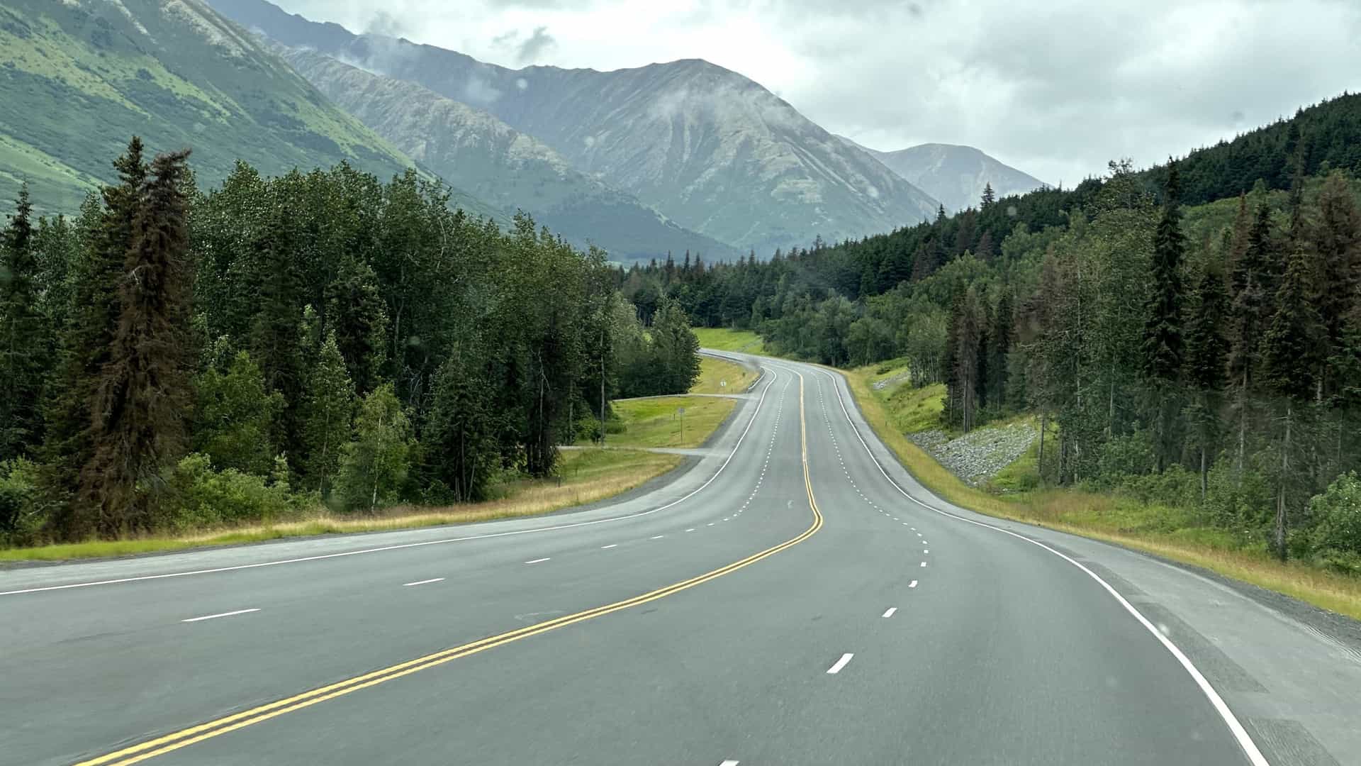 A bright, summertime photograph of a scenic highway in Alaska, possibly the Alaska Highway or Seward Highway, curving through a vast wilderness. The paved road stretches into the distance, flanked by dense green forests and bordered by a wide riverbed or glacial outwash plain. Towering, snow capped mountains dominate the horizon under a bright blue sky.