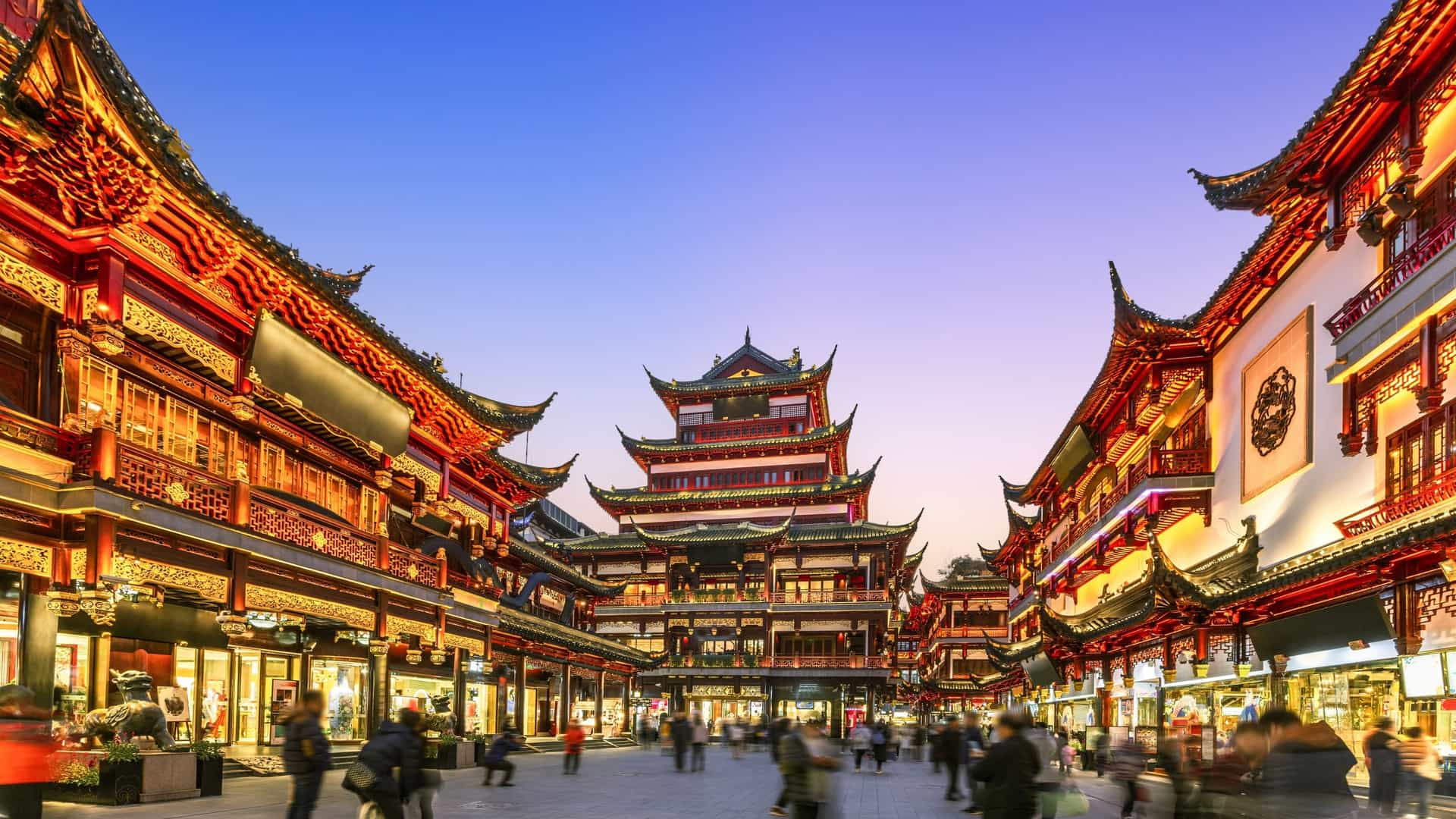A magical night view of the illuminated Yu Garden (Yuyuan Garden) in Shanghai, featuring traditional Chinese pavilions and buildings with curved eaves, brightly lit by numerous red and yellow lanterns and reflected in a tranquil pond or waterway.