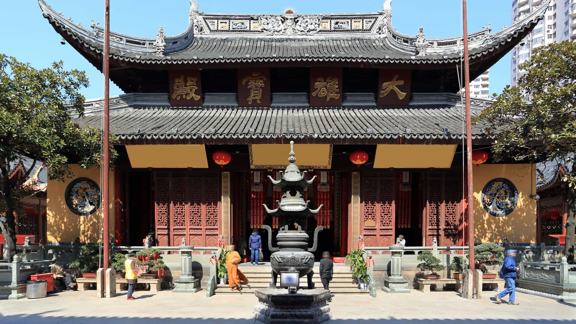 A serene view of the Jade Buddha Temple in Shanghai, a traditional Chinese Buddhist structure featuring classic curved tile roofs and deep brown or reddish wooden architecture, set amidst a peaceful courtyard in the bustling city.
