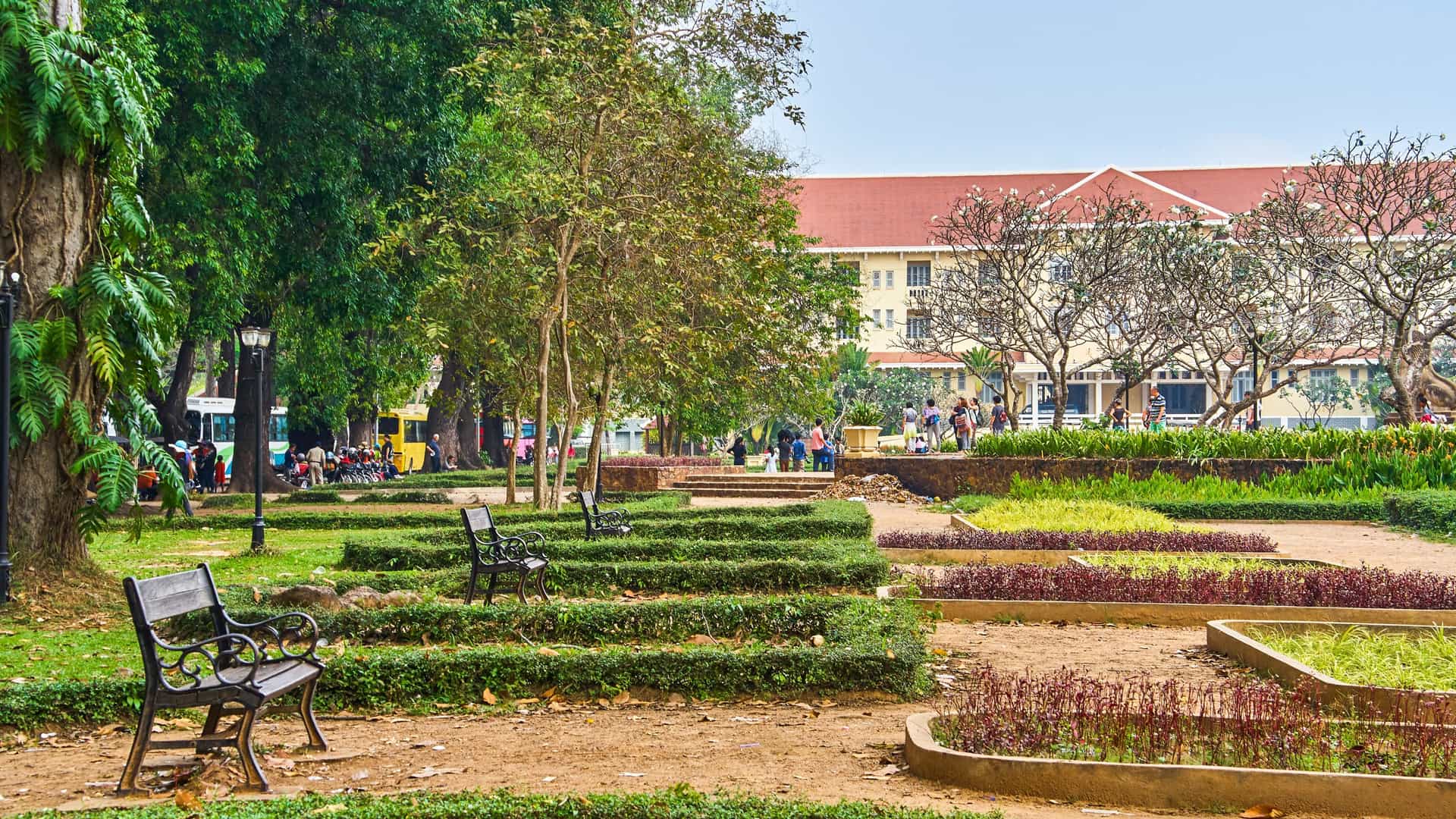 A serene view of the Royal Independence Gardens in Siem Reap, Cambodia, featuring manicured green grass, tall tropical trees, and a decorative, stone statue in the distance, all under a blue sky.