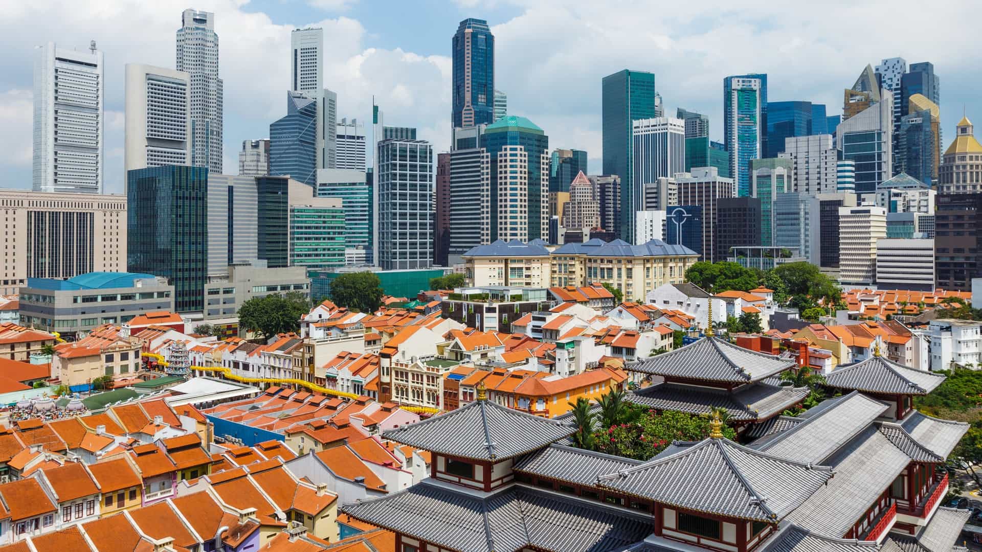 A view of Singapore's Chinatown showing a row of colorful, traditional, low rise shophouses in the foreground, sharply contrasted by the modern, towering glass skyscrapers of the Central Business District (CBD) financial skyline in the background.