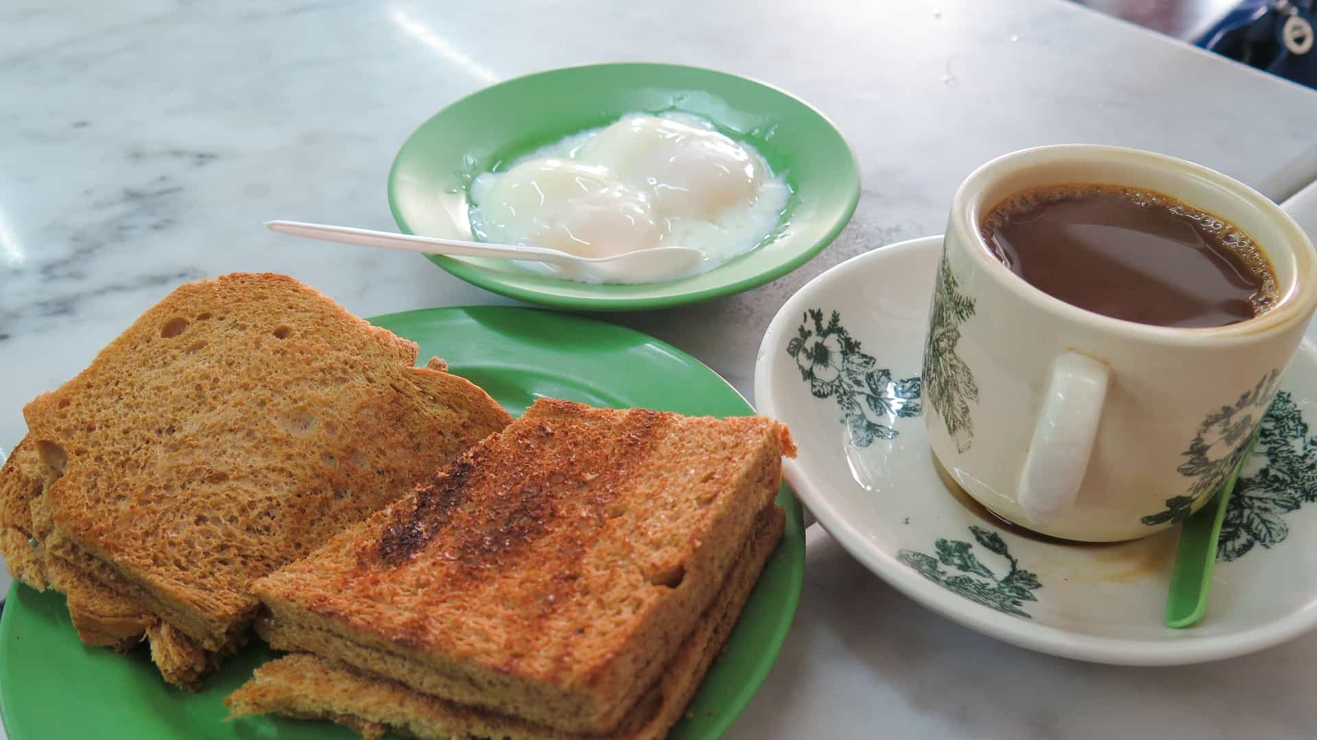 A classic Singaporean breakfast set, often called Kopitiam breakfast, consisting of three items arranged on a wooden table: a plate of kaya toast (two slices of toasted bread with butter and coconut jam), a small bowl containing two soft boiled eggs, and a cup of hot, traditional local coffee (kopi) served in a floral patterned porcelain cup and saucer.