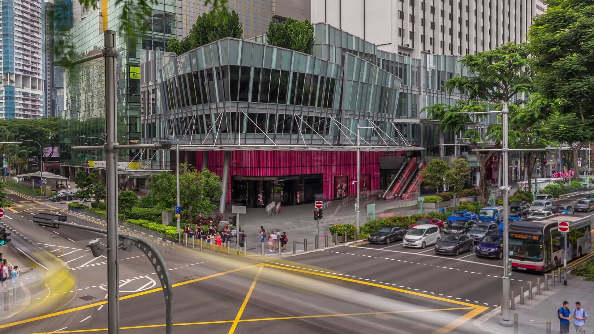 An aerial view of a busy intersection on Orchard Road, Singapore's main shopping district. The wide street is lined with modern high rise buildings and shopping malls, while numerous pedestrians cross the road at the crosswalk.