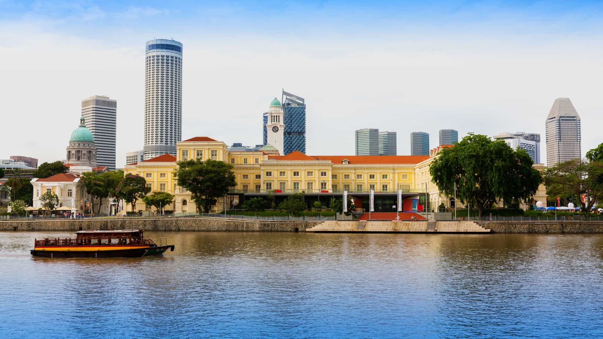 A traditional 'bumboat' river cruise vessel is sailing on the Singapore River in the daytime. The boat is carrying tourists and is cruising past the brightly colored, restored shophouses and modern, glass fronted buildings of Clarke Quay in the background.