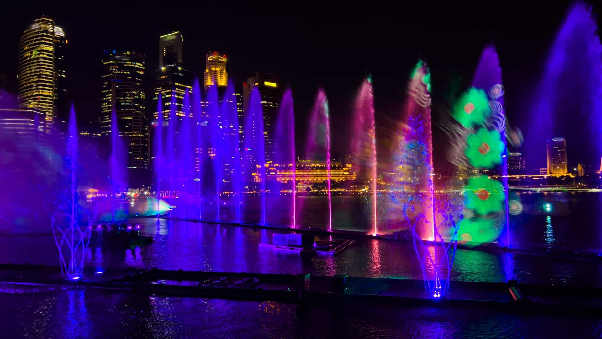 A dramatic night view of the "Spectra" Light and Water Show on the Marina Bay Sands waterfront in Singapore. Multi colored laser beams in red, green, and blue shoot across the sky and water. Fountains of water are lit up with bright colors, creating a dynamic visual display. In the background, the brilliantly illuminated modern skyscrapers of the Central Business District line the cityscape.