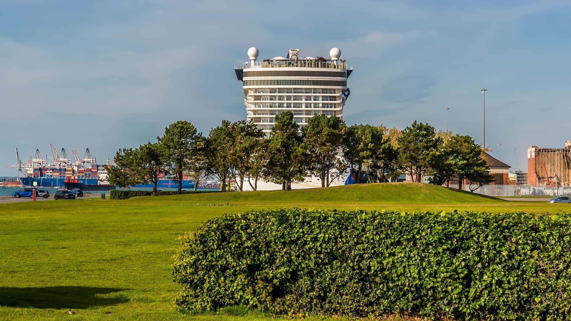 A scenic view across the green expanse of Mayflower Park in Southampton, with the large white hull of a docked cruise ship visible beyond the waterfront. The foreground features grass and trees, framing the massive vessel and the urban docks in the background.