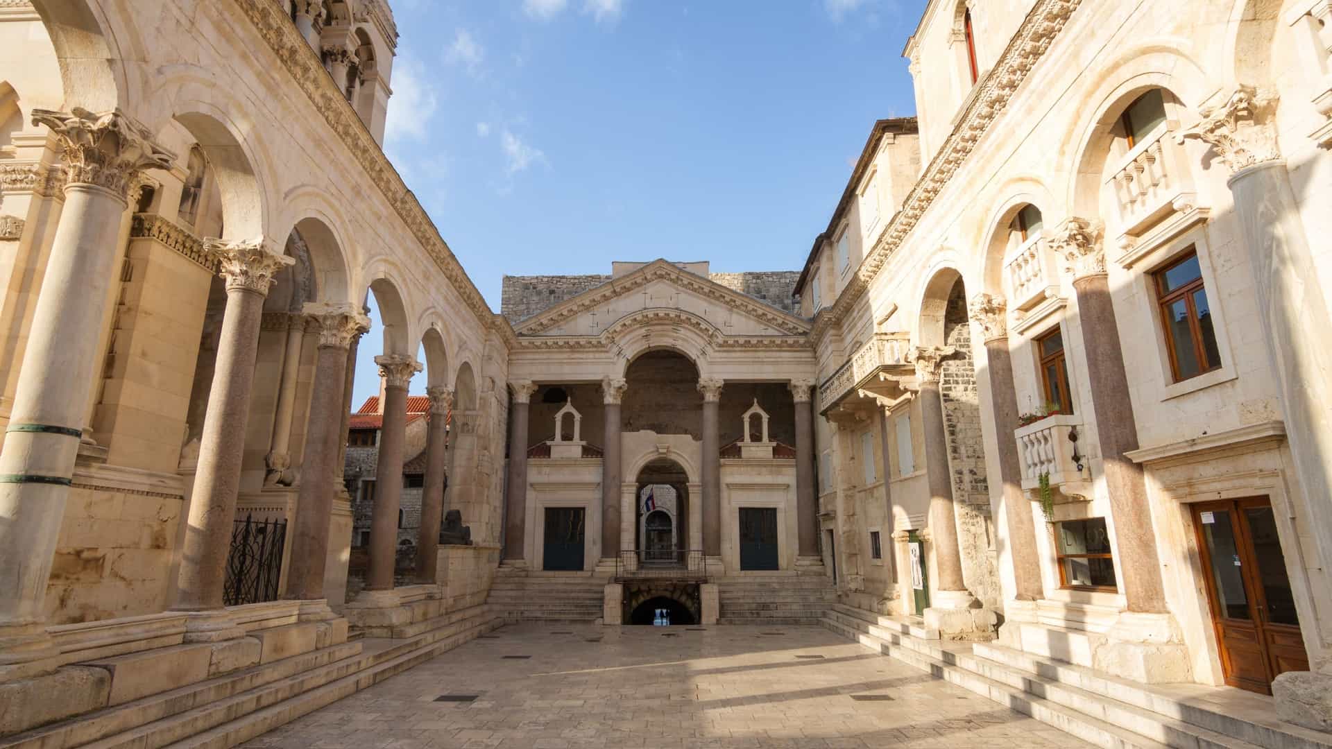A stunning nighttime view of the old, historical Diocletian's Palace in Split, Croatia. The image focuses on the ancient Roman architecture, likely highlighting an illuminated section such as the Peristyle or a facade, with warm lighting contrasting against the dark sky.