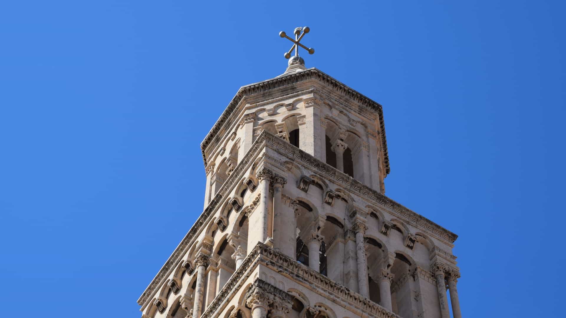 A close up photograph of the upper section of the Bell Tower of the Cathedral of Saint Domnius in Split, Croatia. The image highlights the detailed stone arches, windows, and sculptural elements of the Romanesque structure against the sky, emphasizing the tower's height and historical craftsmanship within Diocletian's Palace.