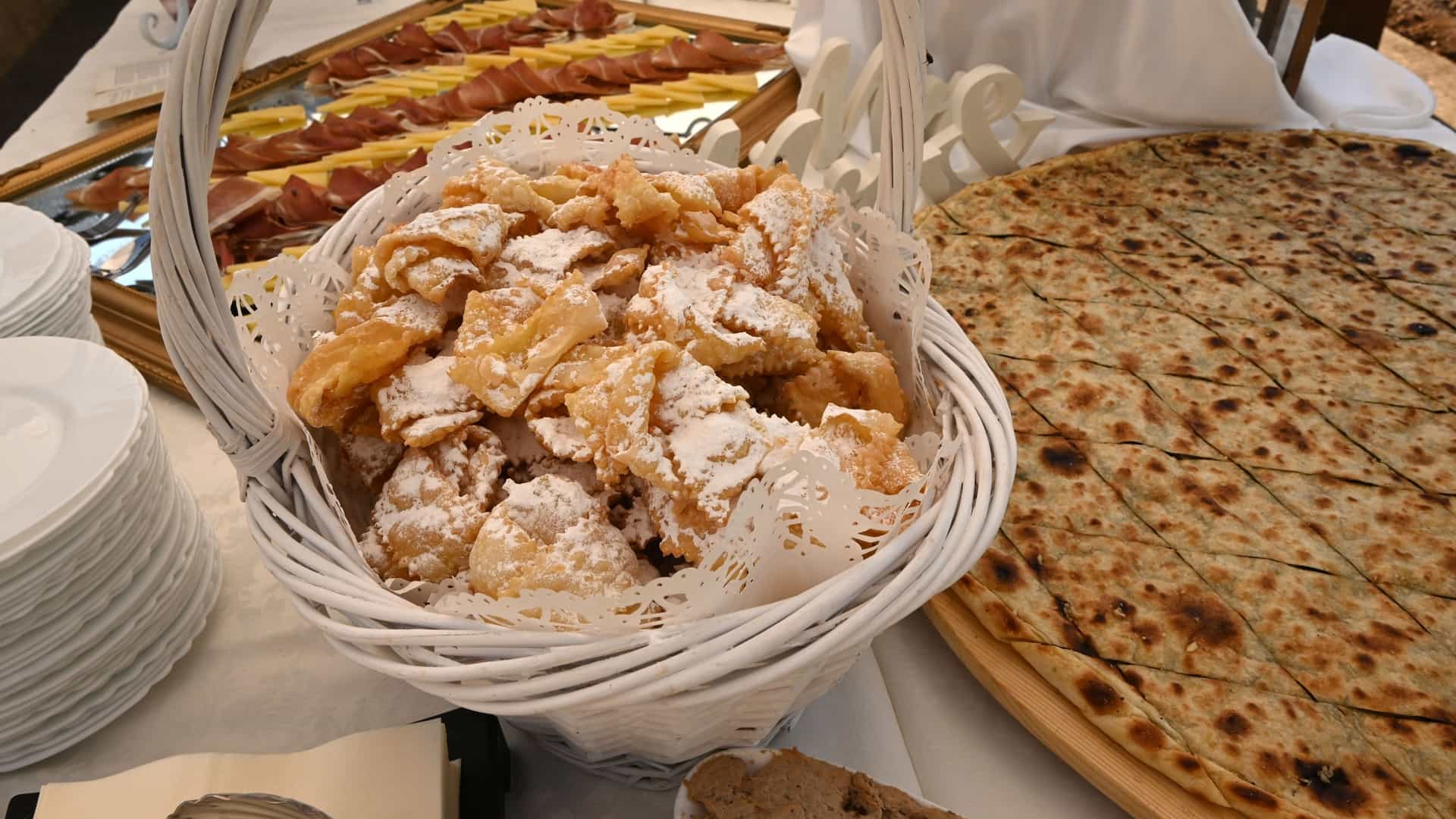 A close up view of a pile of freshly made Kroštule, a traditional Croatian deep fried pastry. The light, golden yellow, ribbon shaped pastries are twisted into knots and generously dusted with white powdered sugar, presented on a plate or in a basket.