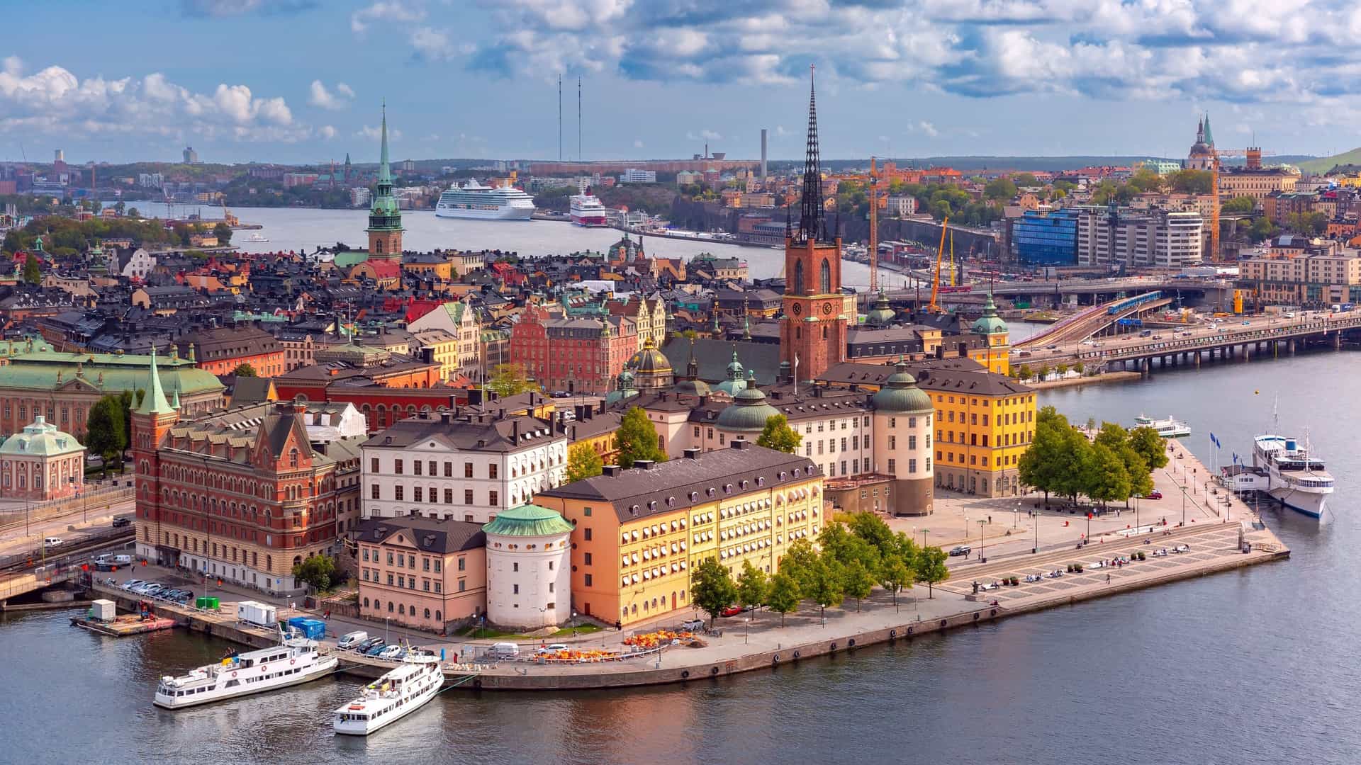 An aerial panoramic view of Gamla Stan, the Old Town of Stockholm, Sweden, featuring a dense cluster of colorful historic buildings with peaked red tiled rooftops, church spires, and a prominent waterway in the foreground under a bright sky.