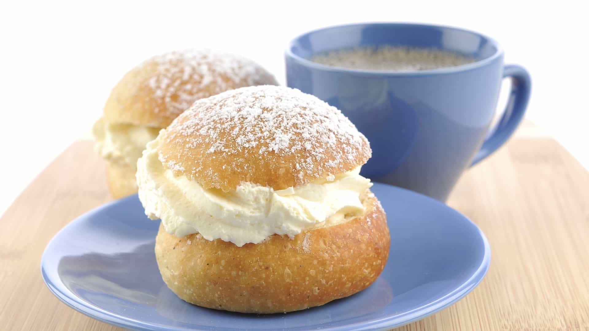A close up shot of a traditional Swedish Semla pastry, or Shrove Bun. The round, cardamom spiced wheat bun is sliced, filled with a rich layer of almond paste and a generous swirl of whipped cream, and finished with the pastry's triangular "lid" dusted with a coating of powdered sugar.