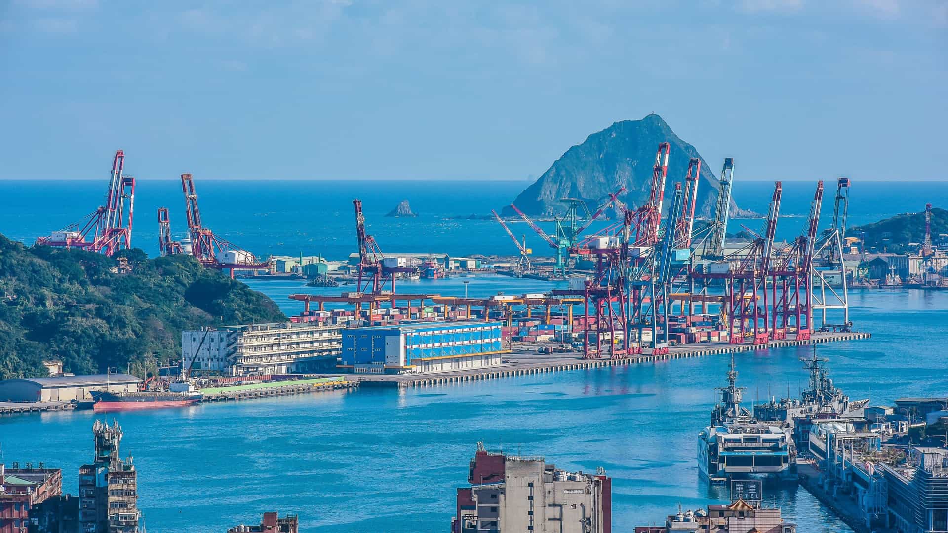 A wide panoramic view of Keelung Harbor and the surrounding city of Keelung, Taiwan, taken from the elevated perspective of Shihciouling Fortification. The scene encompasses the commercial port with docked ships and industrial structures, the dense urban area, and the mountainous coastline under a partly cloudy sky.