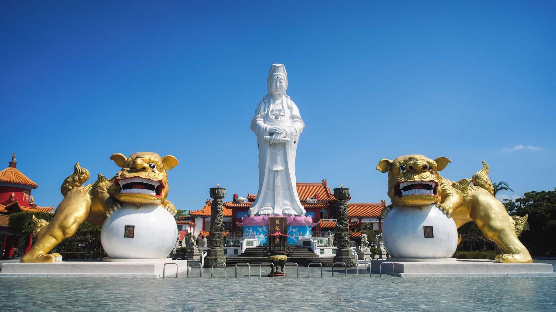 A photograph of the massive, white Statue of Guanyin (Goddess of Mercy) located in Zhongzheng Park in Keelung, Taiwan. The towering statue stands on a pedestal in front of a temple building, overlooking the city and the harbor. The goddess is depicted in a peaceful stance, and the statue is flanked by two large, golden lion statues at its base.