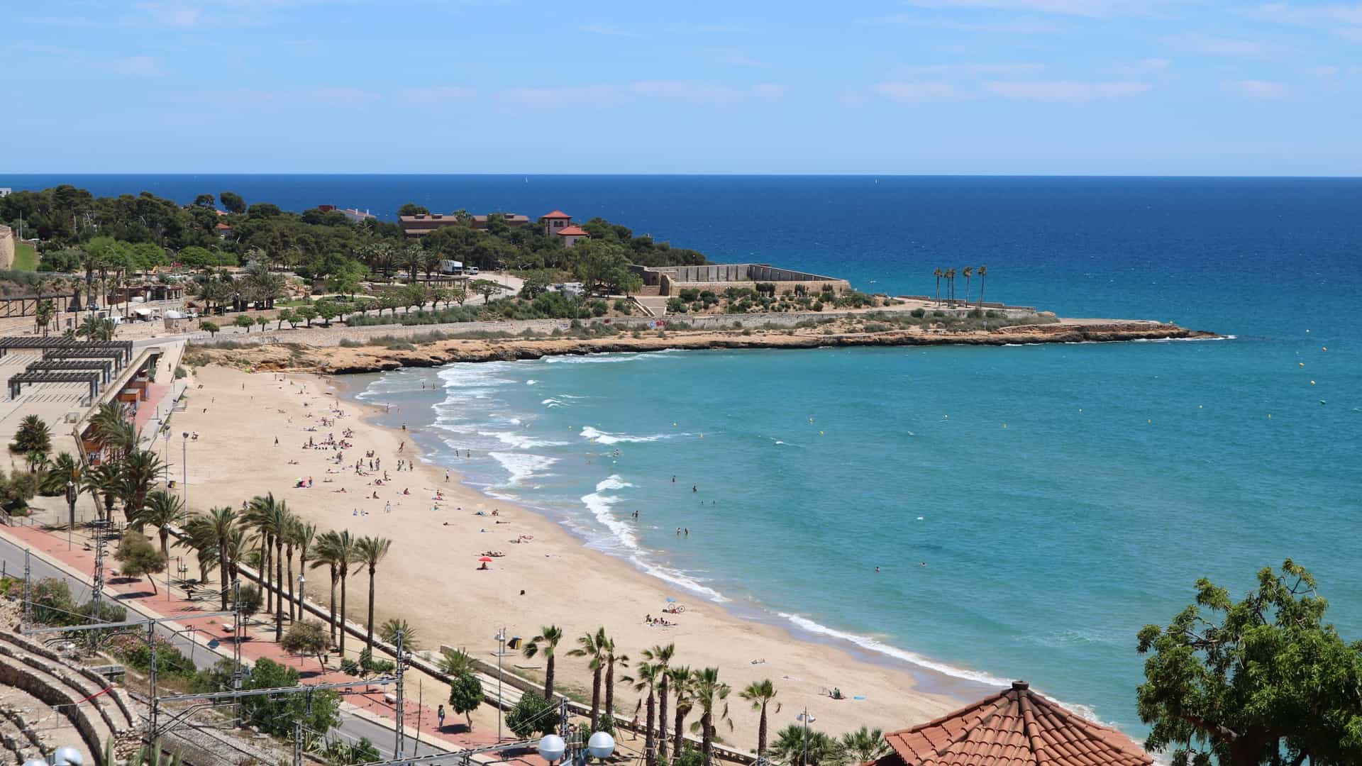 A view of Platja del Miracle (Miracle Beach) in Tarragona, Spain, showing a wide, long stretch of fine golden sand bordering the clear blue Mediterranean Sea. The urban beach is backed by a promenade, with a set of railroad tracks separating the beach area from the city's buildings in the background.