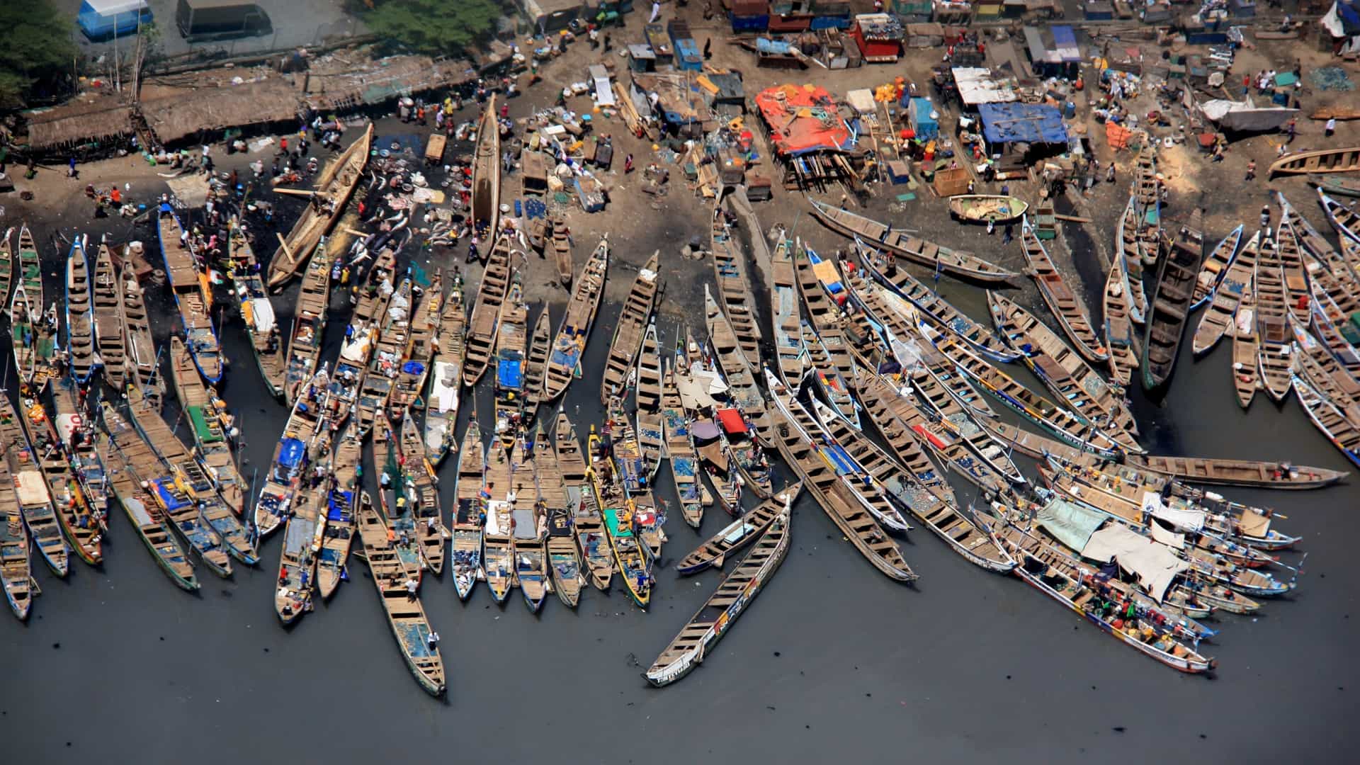 A view of traditional wooden fishing boats, painted in vibrant colors, lined up on a sandy beach in Accra, Ghana. The boats, some with crews of fishermen nearby, are typical of West African coastal communities. The scene suggests a busy fishing harbor or village along the Gulf of Guinea.