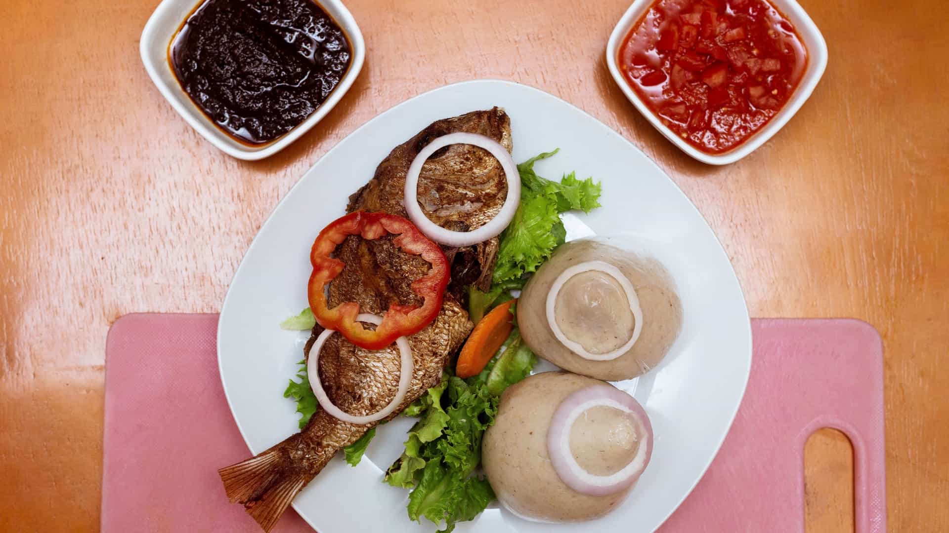 A close up, overhead photo of a plate of traditional Ghanaian cuisine featuring a large portion of Banku (a smooth, whitish ball of fermented corn and cassava dough) served alongside a whole, seasoned fried tilapia fish. The dish is garnished with sliced vegetables like tomatoes and onions, and accompanied by a small bowl of spicy dark brown pepper sauce, known as Shito.