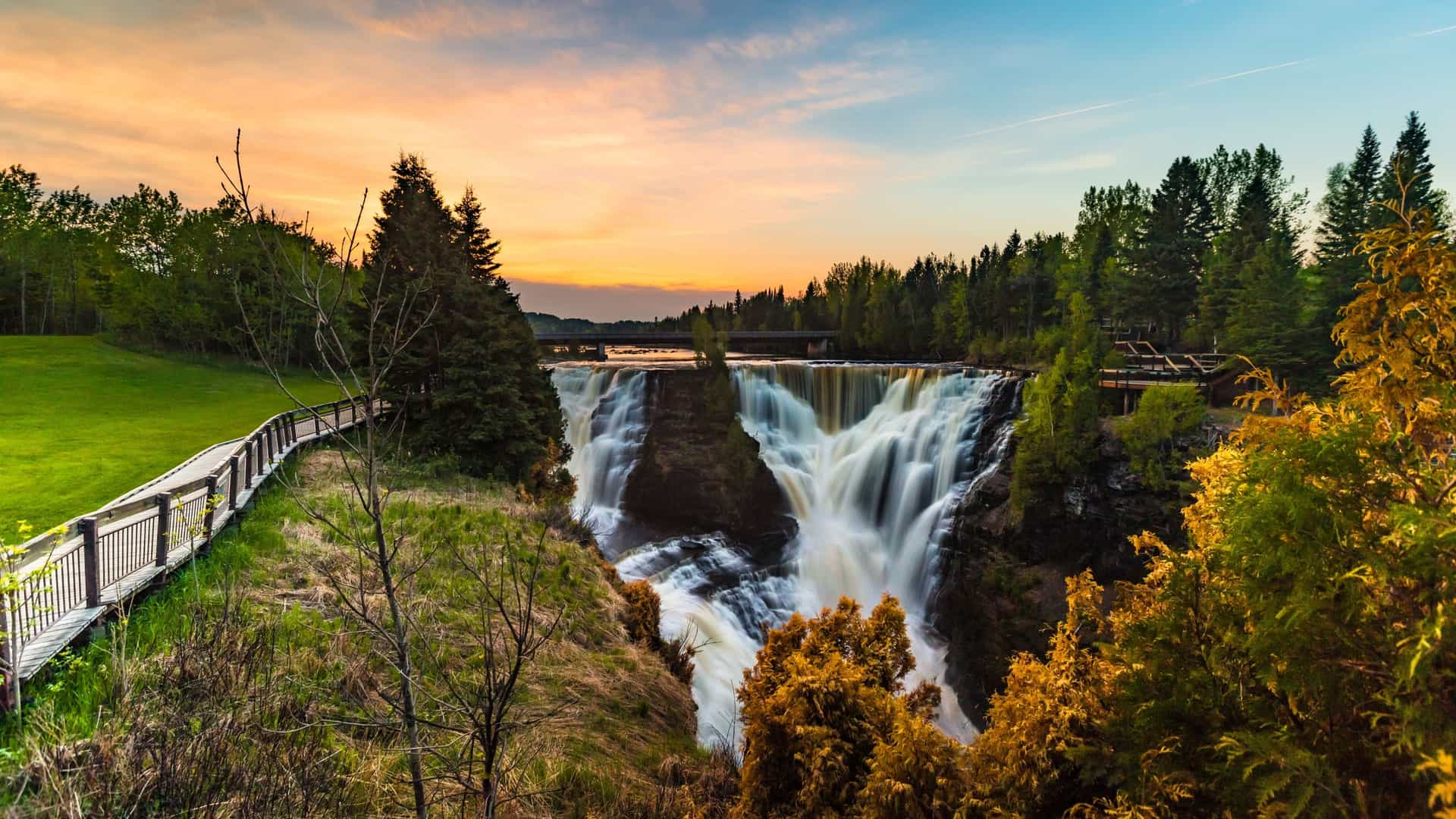 A wide angle view of Kakabeka Falls, known as the "Niagara of the North," where the Kaministiquia River plunges approximately 40 meters (130 feet) over a cliff face and into a rocky gorge. The powerful white water is framed by a dense green forest in Kakabeka Falls Provincial Park, Northern Ontario.