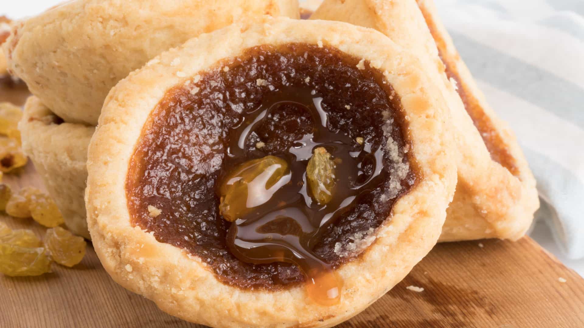 A close up, macro shot of a single butter tart with a flaky pastry crust. The tart is filled with a sweet, gooey, amber colored syrup and features a couple of raisins floating on top. Other tarts are slightly visible and stacked behind it on a wooden board.