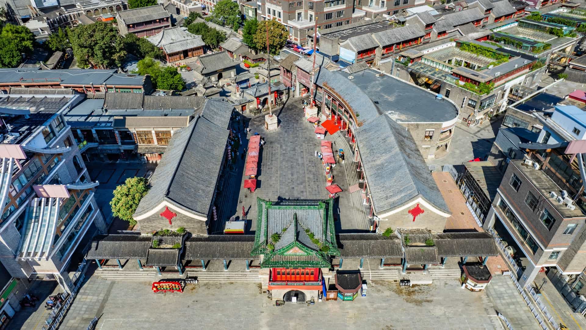 An aerial photograph capturing the Tianjin Ancient Culture Street (Gu Wenhua Jie) in Tianjin, China. The image shows a long pedestrian street lined with traditional Chinese buildings featuring classic multi tiered, tiled roofs and colorful facades, contrasting with the modern high rise buildings in the distance. The central Tianhou Temple is visible in the middle of the historical buildings. The scene is captured during the day under bright, clear conditions.
