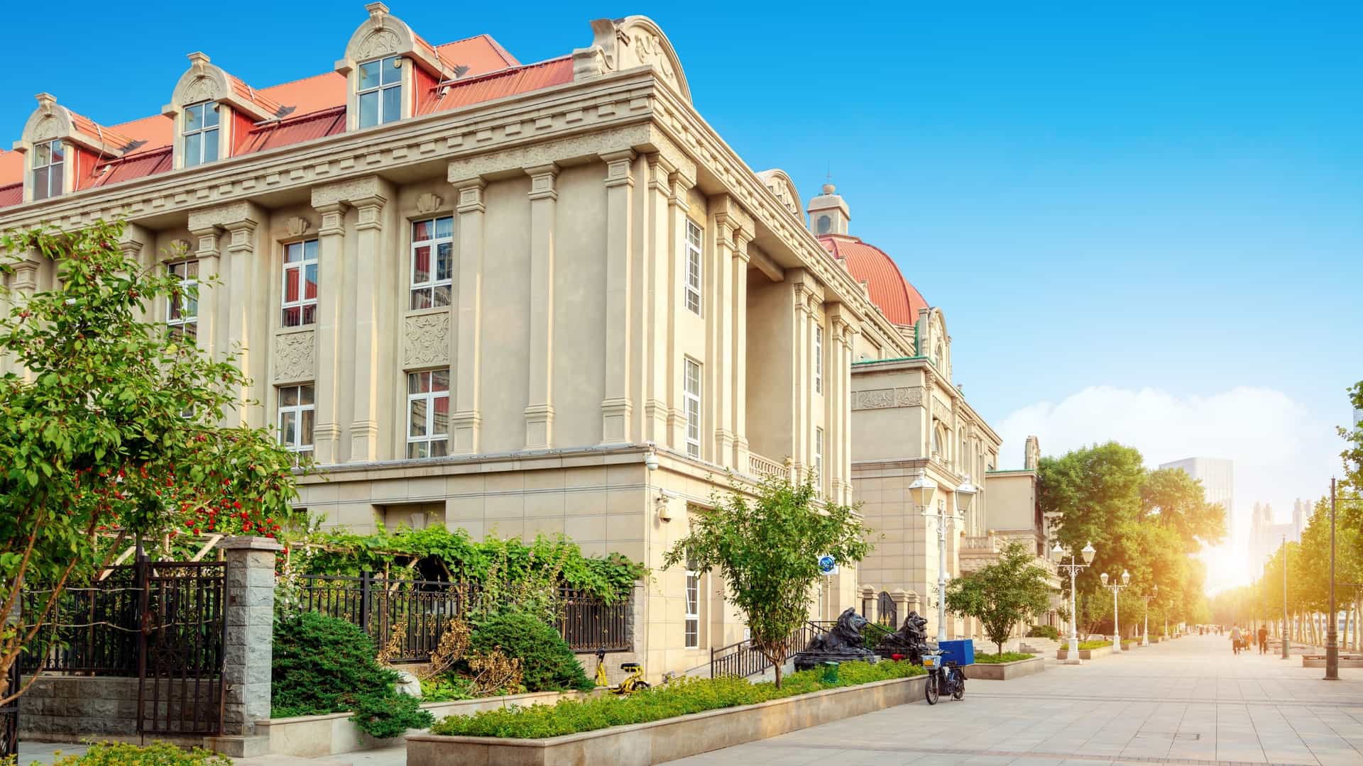 A large, historic, Italian style building with a light beige facade, prominent columns, and a terracotta colored mansard roof, located on a wide paved pedestrian boulevard, likely in the Tianjin Italian Style Street in China. The building is bordered by lush green landscaping and a black metal fence, with the boulevard and streetlights stretching into the distance under a bright sun flare.