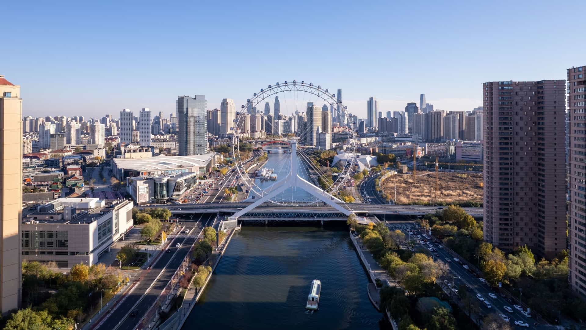 An aerial shot of the Tianjin Eye, a giant Ferris wheel, positioned over the Yongle Bridge which crosses the Haihe River. The modern skyline of Tianjin, China, with numerous skyscrapers, is visible across the city, all under a clear sky.