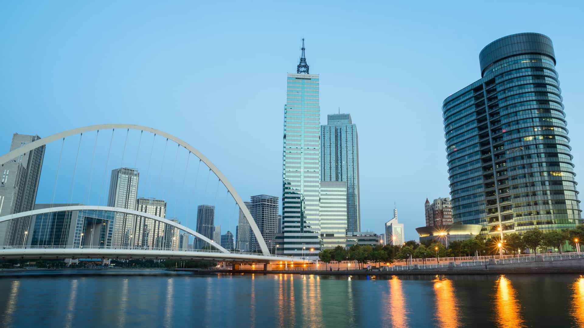 The downtown skyline of Tianjin, China, featuring modern high rise buildings and skyscrapers, with the Haihe River flowing in the foreground. The city lights are illuminated, reflecting off the river at dusk or nighttime.