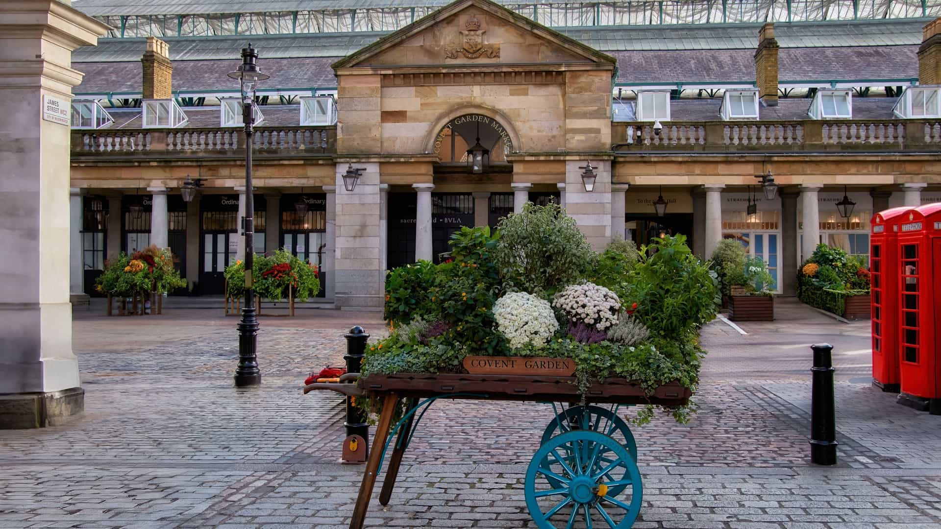 A charming wooden floral cart in Covent Garden, London, filled with colorful flowers and plants. The cart is typically positioned against a backdrop of historic architecture, sometimes including the famous red telephone boxes, as a tribute to the area's history as a flower market.