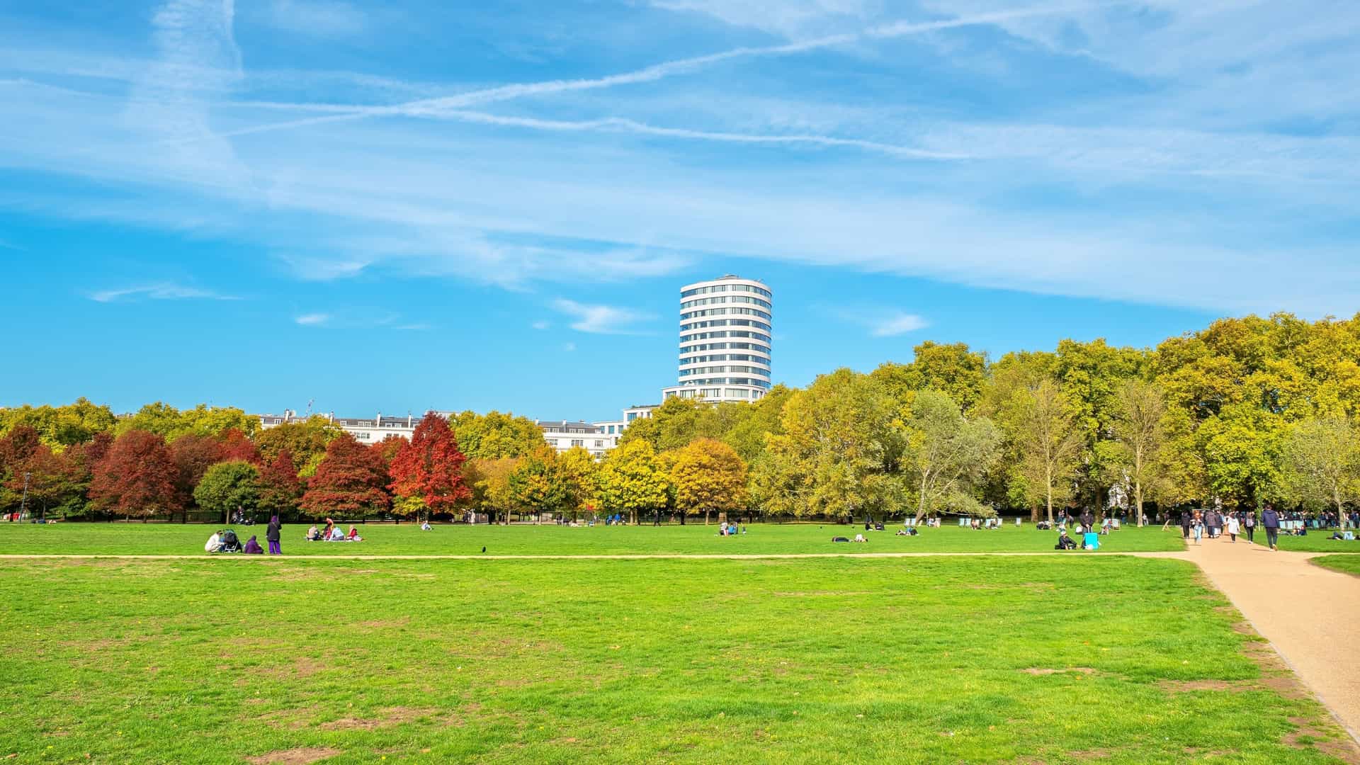 A scenic view of Hyde Park in London during autumn, featuring a path or avenue lined with trees displaying vibrant fall foliage in shades of yellow, gold, and red. The scene often includes fallen leaves on the ground, bright sunlight, and a tranquil atmosphere.
