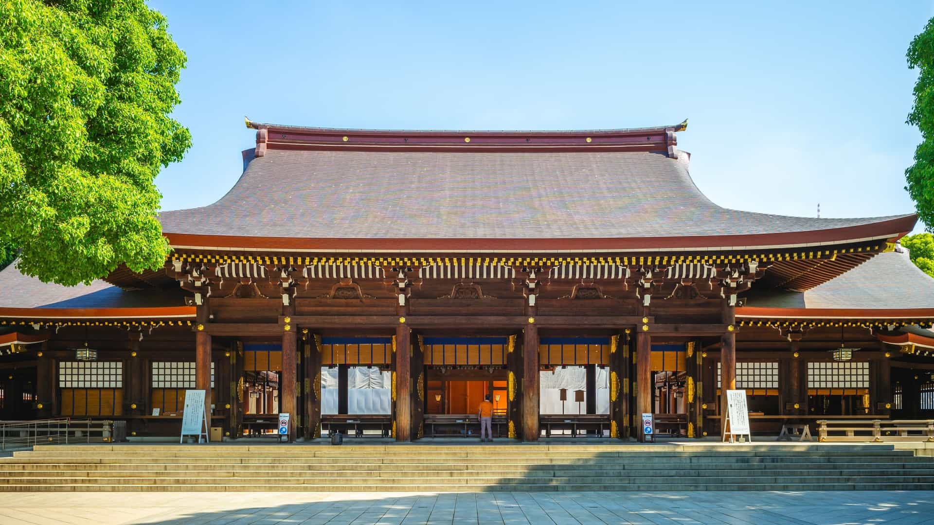 The Main Hall (Honden) of the Meiji Shrine in the Shibuya district of Tokyo, a traditional Japanese Shinto building constructed of unpainted wood and featuring a distinctive copper roof. The building is situated behind a wooden gate within a large, raked gravel courtyard and surrounded by the dense green forest of the shrine's grounds.