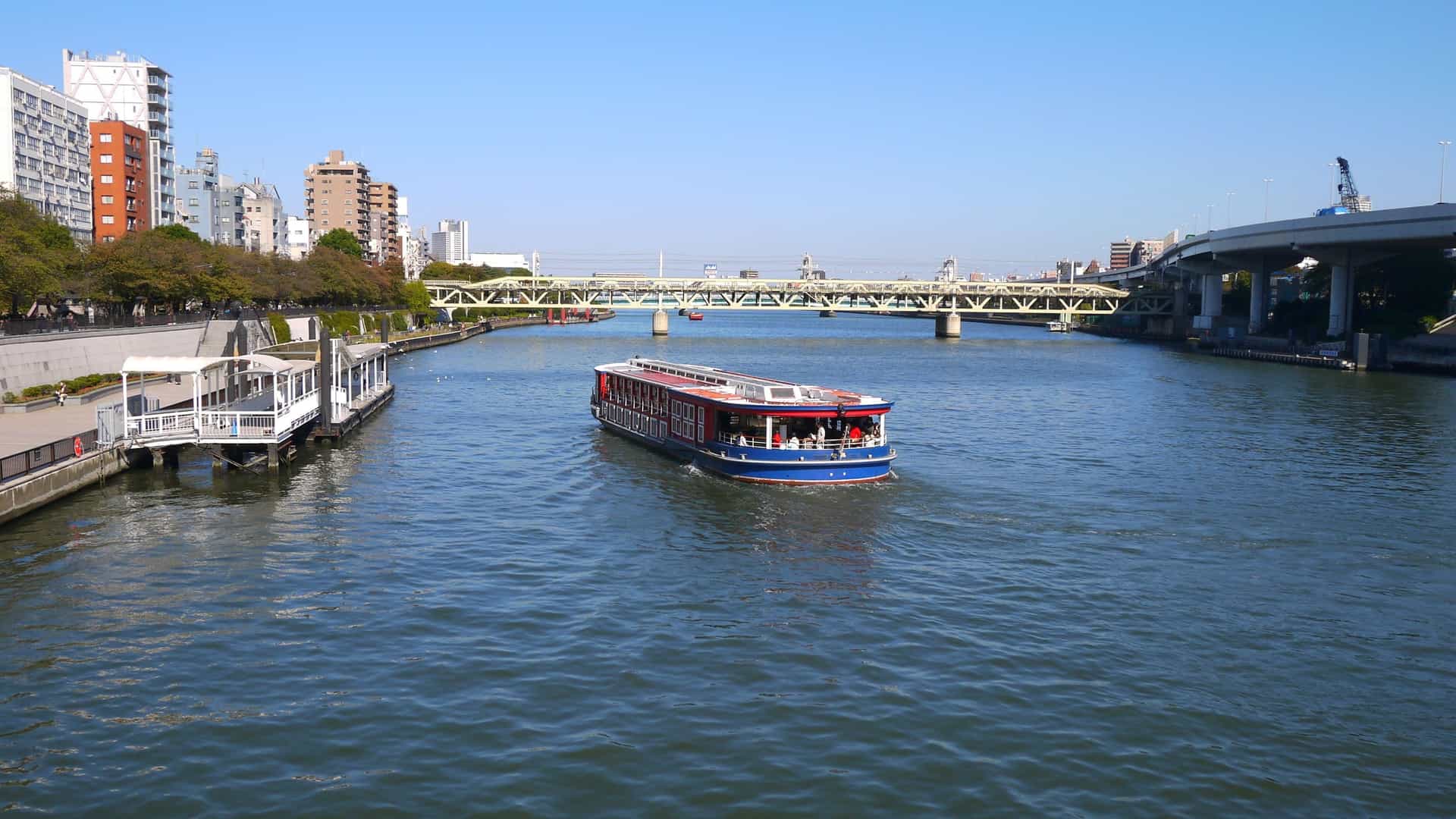 A view of the Sumida River in Tokyo, Japan, with a modern cityscape on its banks. Prominently featured are the towering Tokyo Skytree and the golden, flame shaped structure of the Asahi Beer Hall near the river.