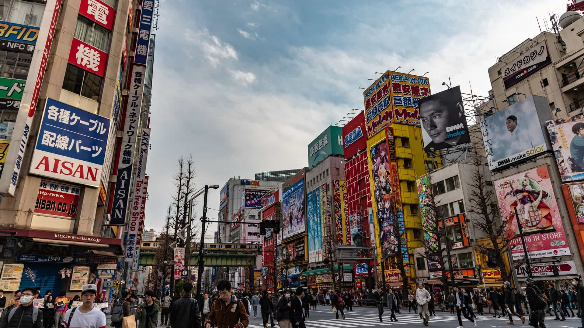 A night view of the bustling streets of Akihabara Electric Town in Tokyo, Japan. The main street is lined with multi story buildings covered in large, colorful, illuminated billboards and signs advertising electronics, anime, manga, and video games. Pedestrians are walking on the sidewalk and crossing the street, creating a vibrant, high energy urban scene.