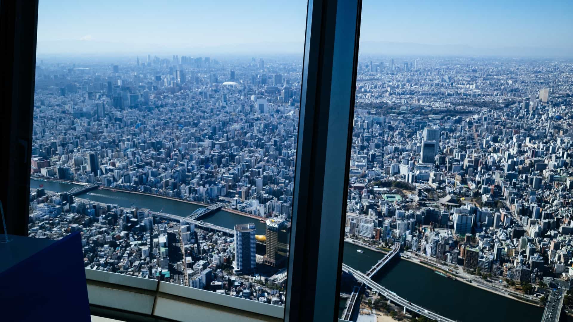 A bright, panoramic daytime aerial view of the Tokyo metropolitan area, captured from the Tokyo Skytree observation deck. The dense urban landscape of skyscrapers, mid rise buildings, and houses stretches out to the horizon under a clear blue sky. In the far distance, a recognizable Mount Fuji is visible on the left, its snowy peak rising above the haze, with a prominent road or highway cutting through the cityscape in the foreground.