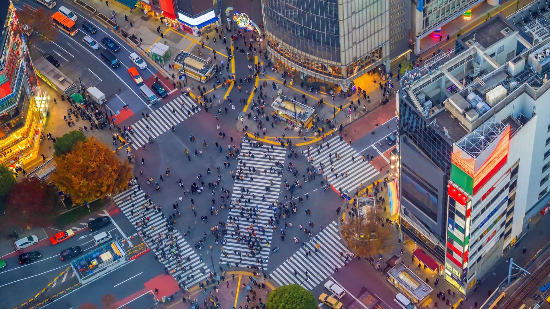 An iconic, high angle night view of the bustling Shibuya Crossing in Tokyo, Japan. The image captures the famous scramble intersection from above, showcasing a dense crowd of pedestrians crossing the street, creating motion blur lines. The intersection is surrounded by tall buildings, whose facades are covered in vibrant, illuminated neon signs and large video screens displaying advertisements, bathing the scene in a kaleidoscope of colors. The overall effect is one of a dense, electric, and perpetually busy urban nightlife scene.