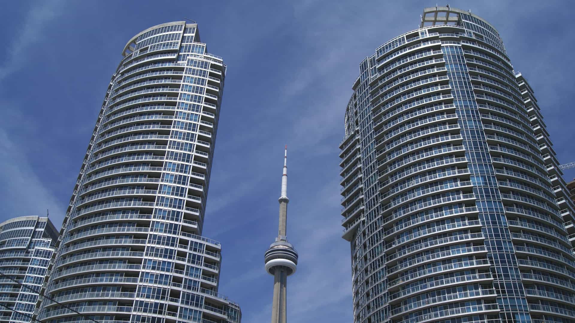A prominent view of the CN Tower in Toronto, Canada, a tall concrete and steel telecommunications and observation tower. The structure's main shaft extends into the sky, crowned by the spherical main pod and topped with a metal antenna, set against a bright, clear sky.