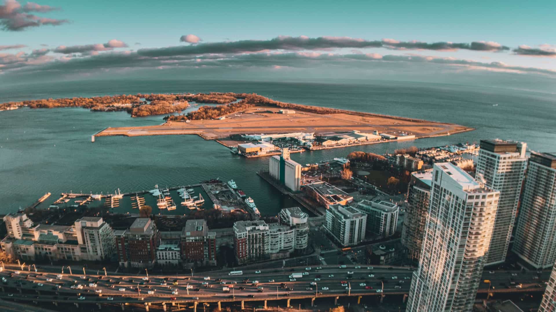 A high angle aerial view captures the dense Toronto skyline dominated by towering skyscrapers and the iconic CN Tower. In the foreground, the city's waterfront is visible, featuring a marina filled with boats on the blue waters of Lake Ontario.
