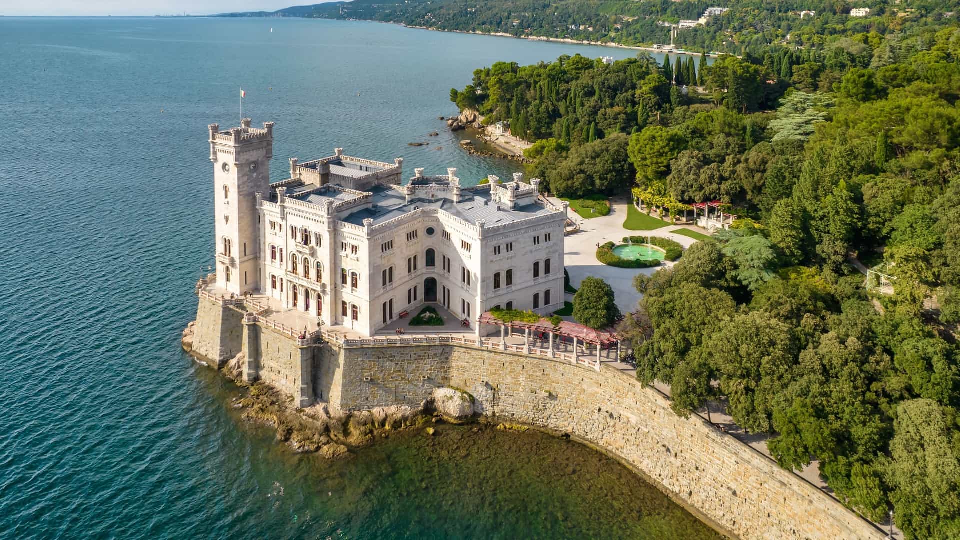 An aerial and panoramic view of the Miramare Castle (Castello di Miramare), a stunning white, 19th century castle with neo Gothic architecture, situated on a rocky promontory on the Gulf of Trieste in Italy. The castle is surrounded by its large, lush green park and the deep blue waters of the Adriatic Sea, showcasing a picturesque and dramatic coastal setting.