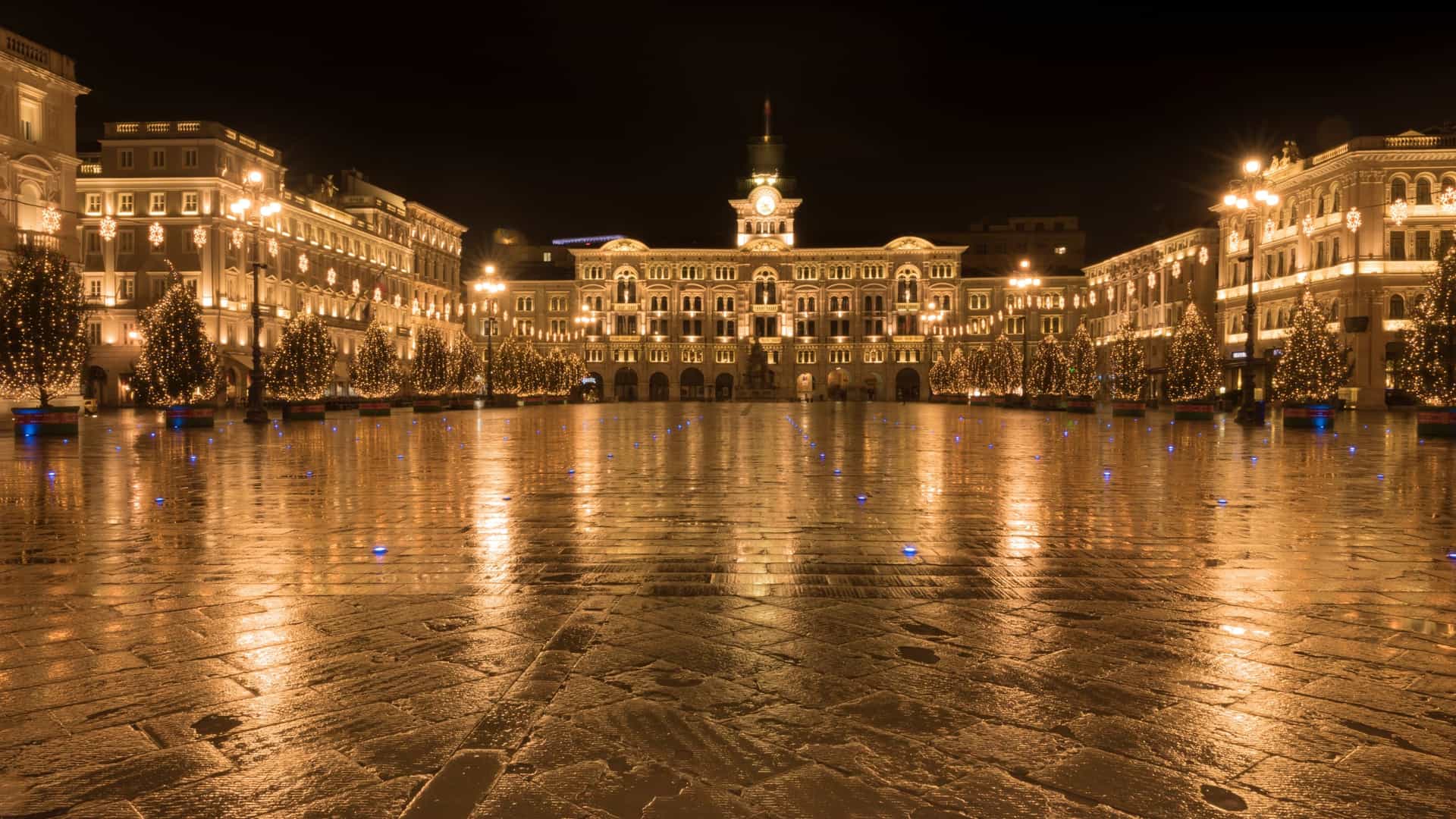 A wide angle view of the Piazza dell'Unità d'Italia (Unity of Italy Square), the grand main square of Trieste, Italy. The square is paved and bordered by imposing, historic neoclassical palaces, including the Palazzo del Municipio (Town Hall) with its central clock tower. In the foreground, the square features the Fountain of the Four Continents, and the background shows the square opening up to the Adriatic Sea.
