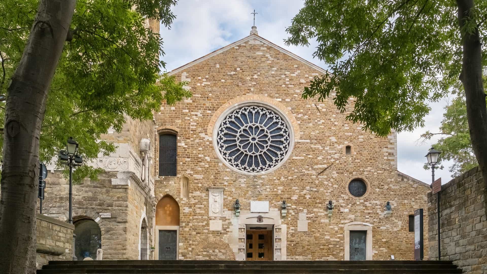 The facade of the Trieste Cathedral (Cattedrale di San Giusto Martire) on the San Giusto hill in Trieste, Italy. The Romanesque Gothic church features an austere stone facade dominated by a large, intricately carved Gothic rose window. To the right stands the massive, squat bell tower, which incorporates the remains of an ancient Roman temple (Propylaeum).