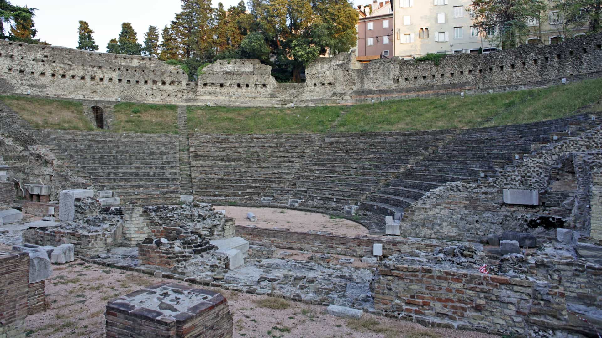 The well preserved ruins of the Roman Theatre (Teatro Romano) in Trieste, Italy. The ancient stone structure, built between the 1st and 2nd centuries AD, shows tiered semi circular seating (the cavea) and the remains of the stage area, all nestled right in the middle of a modern city center at the foot of the San Giusto hill, with contemporary buildings visible beyond the ruins.
