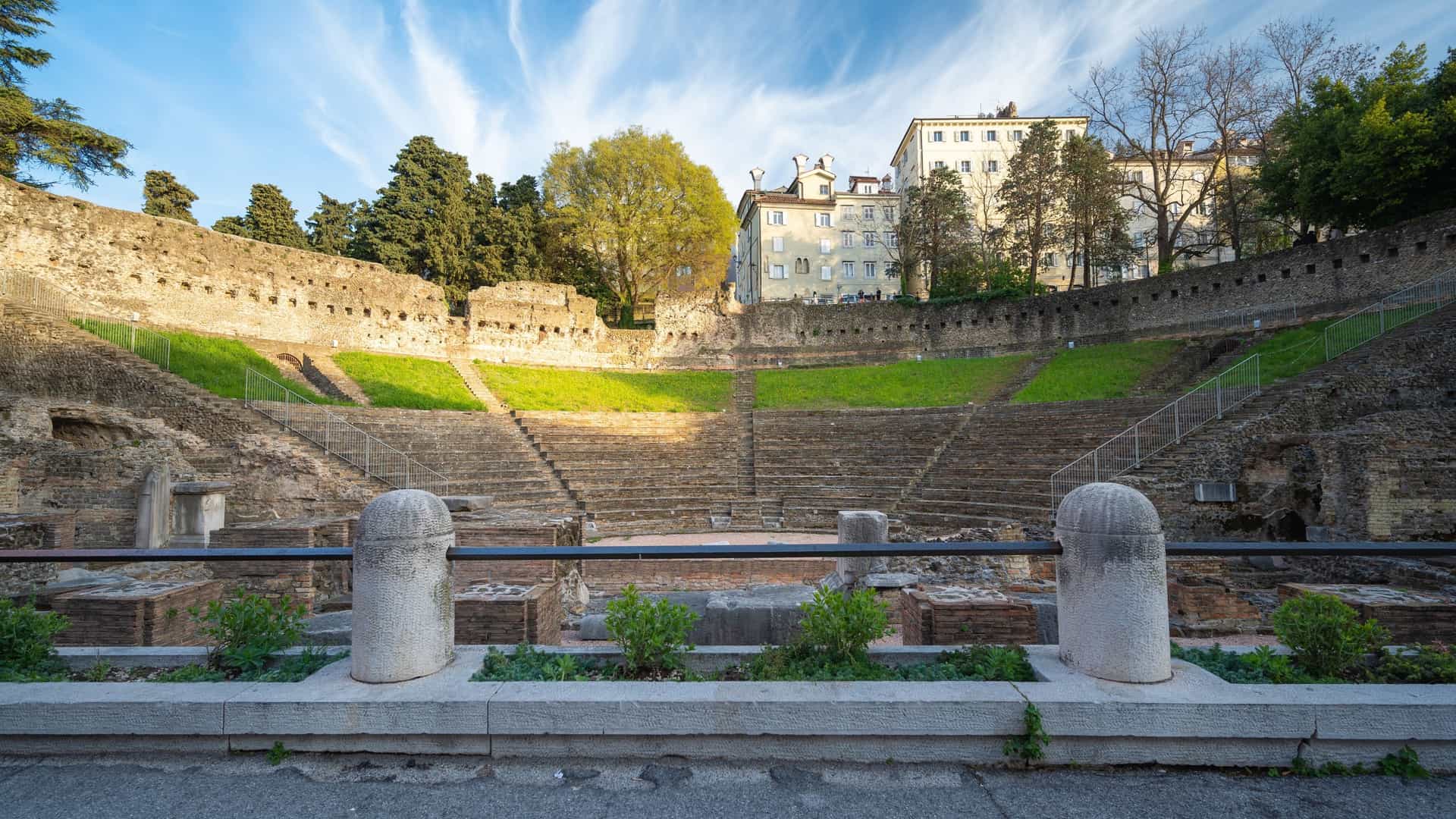 A close up view of the stone and brick ruins of the Roman Theatre (Teatro Romano) in Trieste, Italy, showcasing the well preserved tiered seating (cavea). The ancient Roman structure, dating back to the 1st century AD, is highlighted against the backdrop of modern city buildings, emphasizing the juxtaposition of history and contemporary urban life.