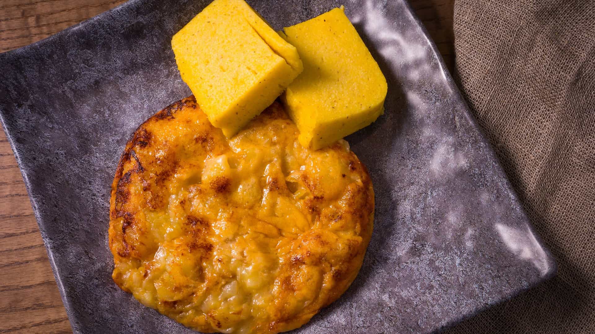 A traditional Friulian dish called Frico, a thick, savory pancake made from pan fried Montasio cheese and potatoes, served on a white plate. The frico has a golden brown, crispy crust and a soft interior. It is typically accompanied by slices of yellow polenta and sometimes garnished with herbs, representing a classic, hearty dish from the Friuli Venezia Giulia region of Italy.