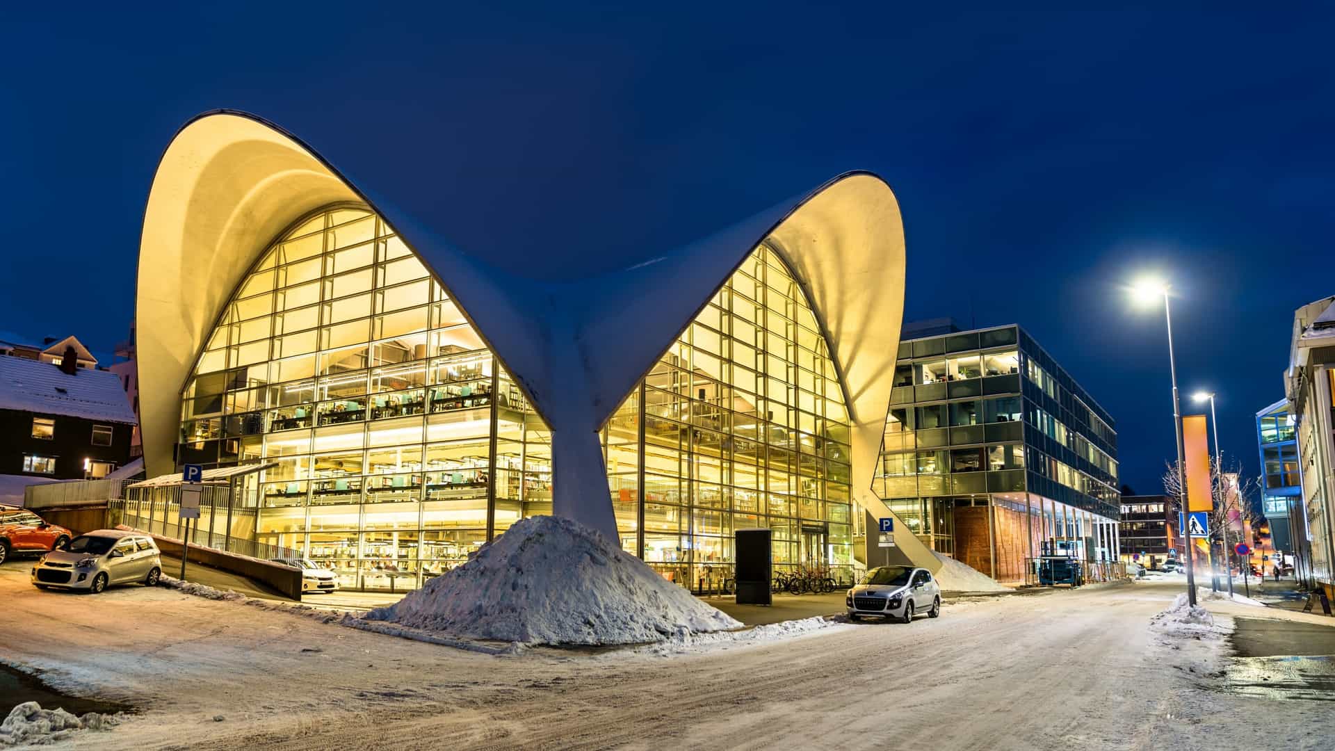 An exterior view of the Tromsø City Library and Archive in Norway, showcasing its striking, modern architecture. The building features a large, flowing, hyperbolic paraboloid roof—the former roof of the Fokus Cinema—supported by a glass facade. The transparent walls reveal the bright, multi level interior, and the library stands prominently in the city center.