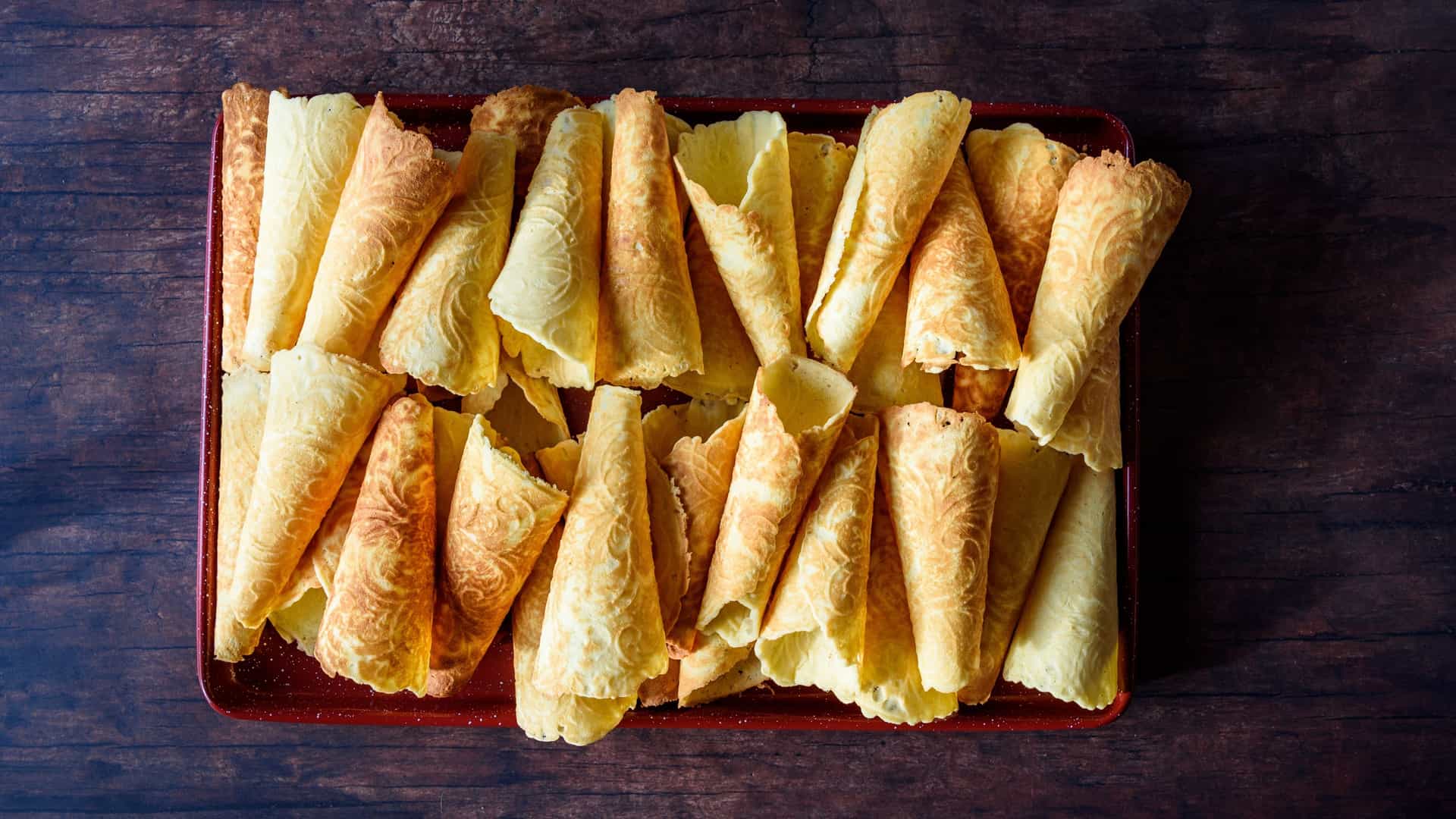 A close up of several traditional Norwegian Krumkake cookies. The cookies are thin, golden brown, and delicately rolled into crisp conical shapes. They are arranged on a bright red serving tray, which is placed on a rustic, dark wooden table.